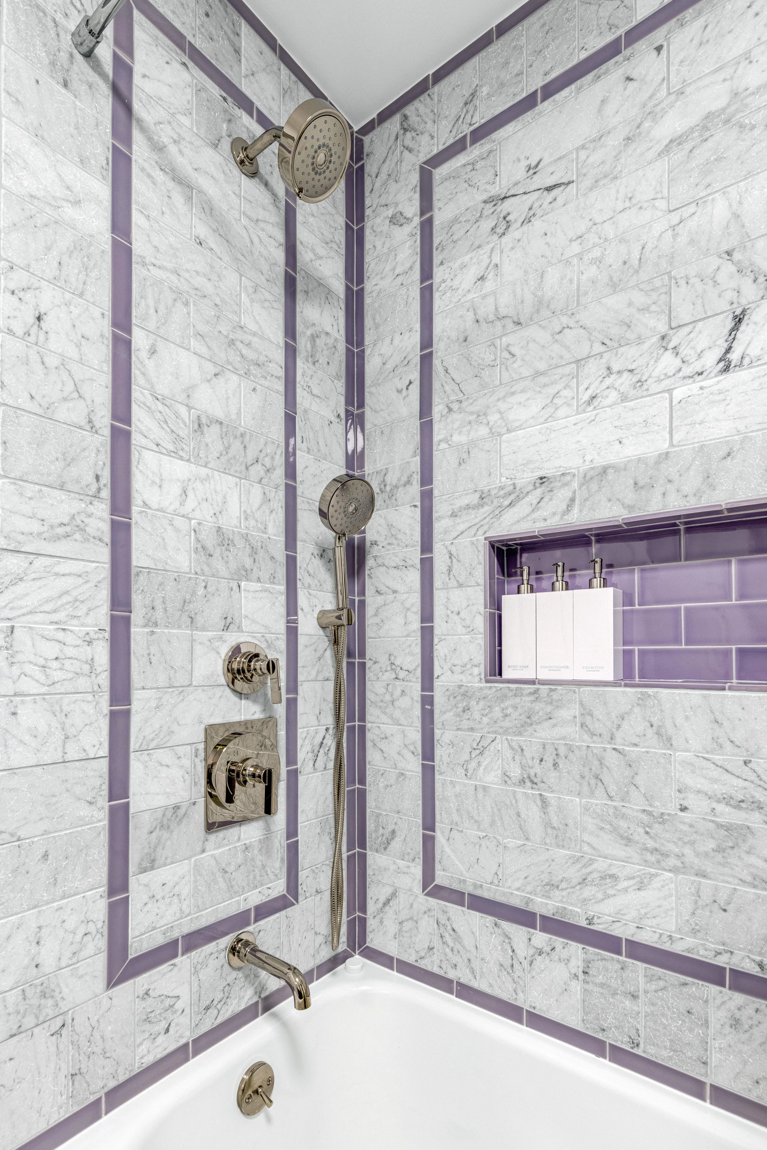 A bathroom shower with purple tile accents, a marble wall, and a built-in shelf with toiletries.