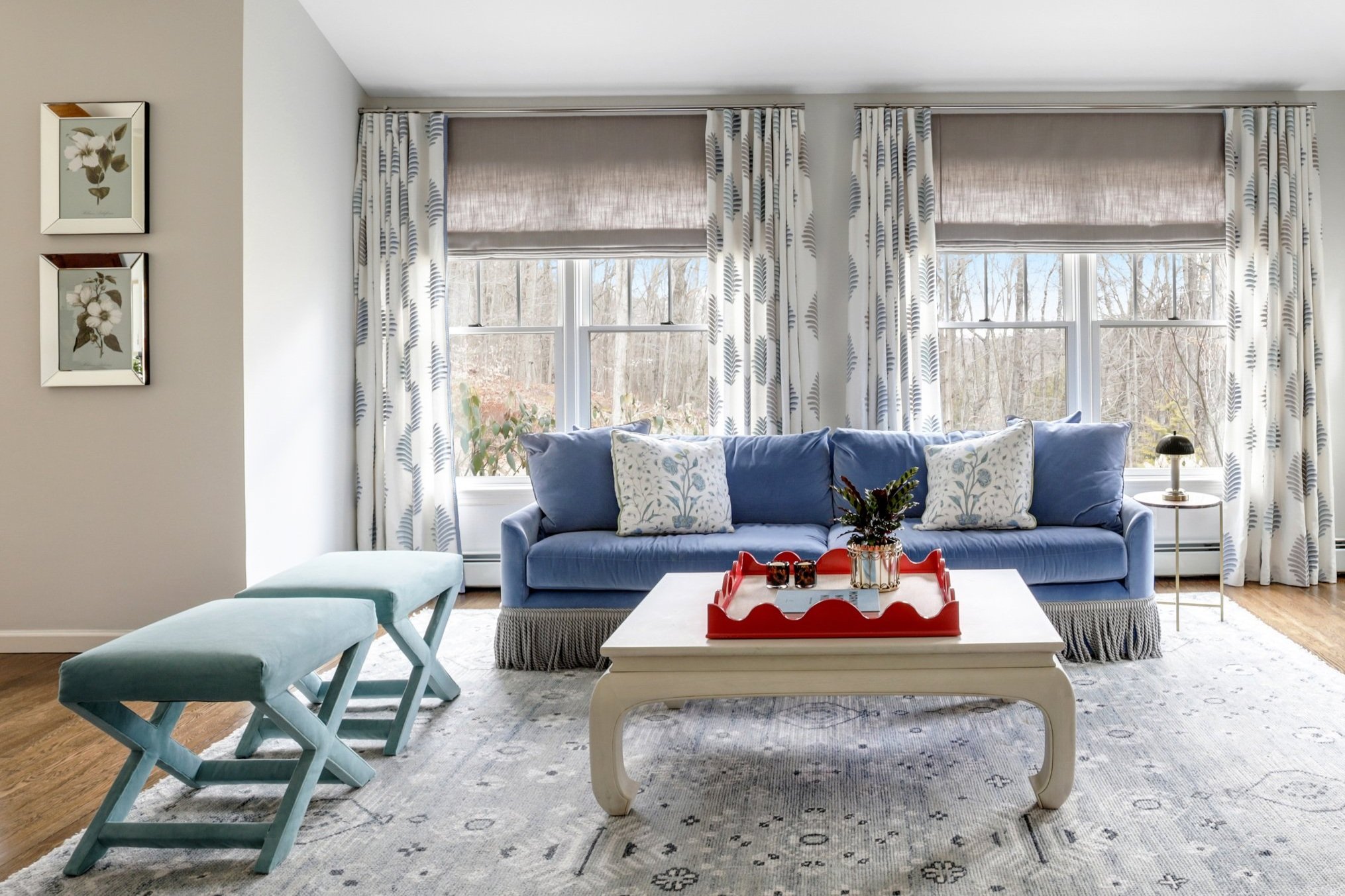 Living room with white walls, large windows with patterned curtains, a blue sofa with decorative throw pillows, a white coffee table with a red tray and plant, two light blue benches, a side table with a lamp, and framed floral artwork on the wall.