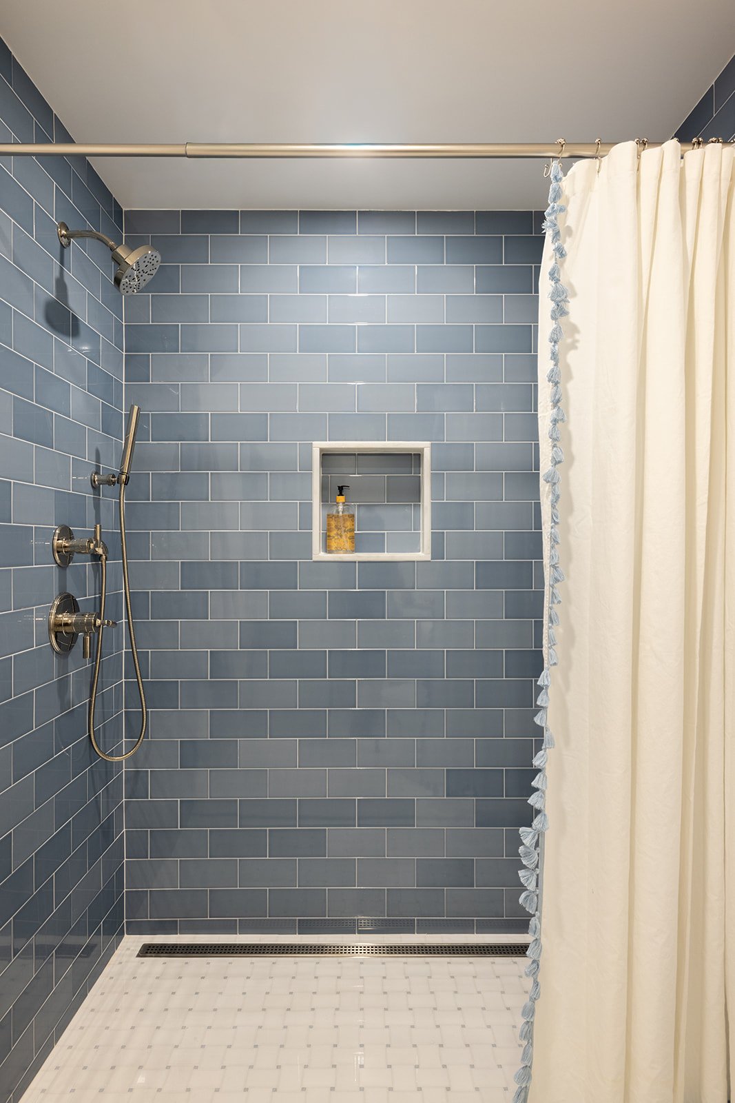 A modern walk-in shower with blue subway tiles, a built-in niche with soap, a rain showerhead, a handheld shower, a curved curtain rod, and a cream shower curtain with blue trim.