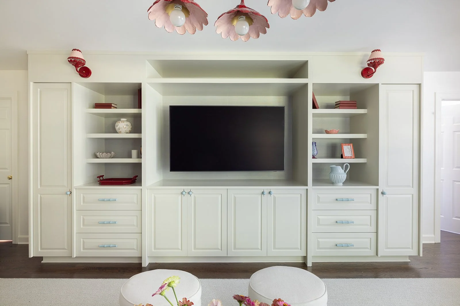 Living room with white built-in shelving unit, a central black flat-screen TV, and decorative items like books, vases, and bowls. Three pink pendant lights hang from the ceiling.