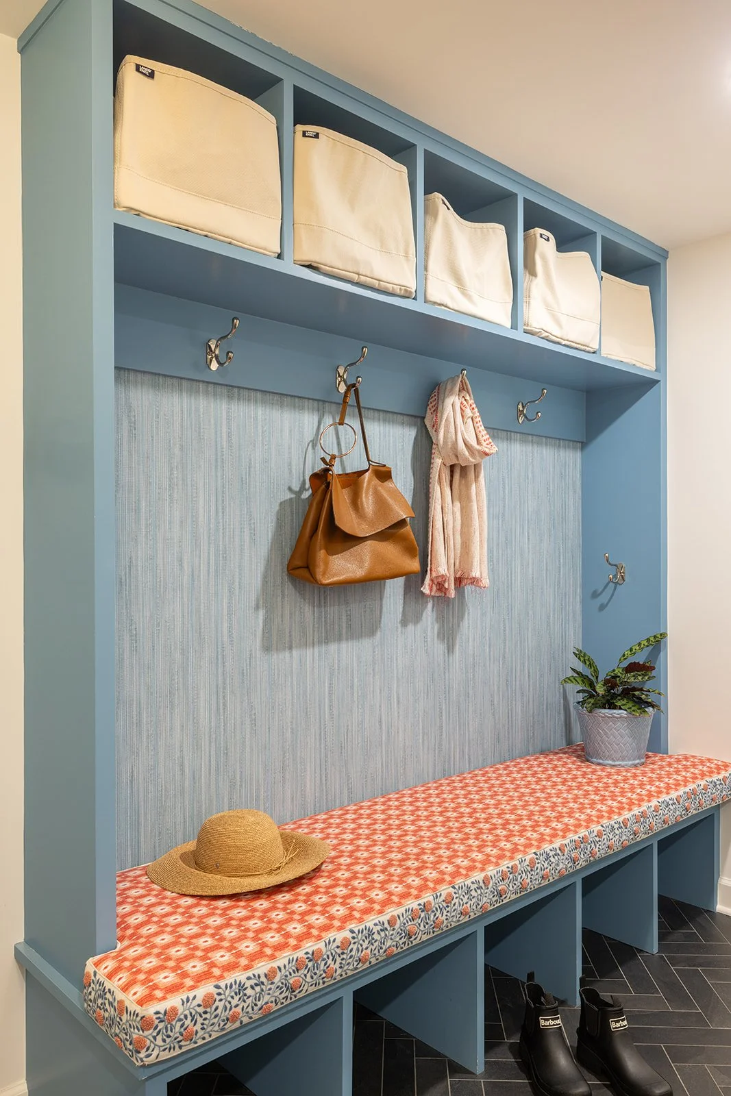 Blue mudroom with hooks holding a brown handbag and pink scarf, a bench with a patterned cushion, a hat and a plant, and storage cubbies with fabric bins above.