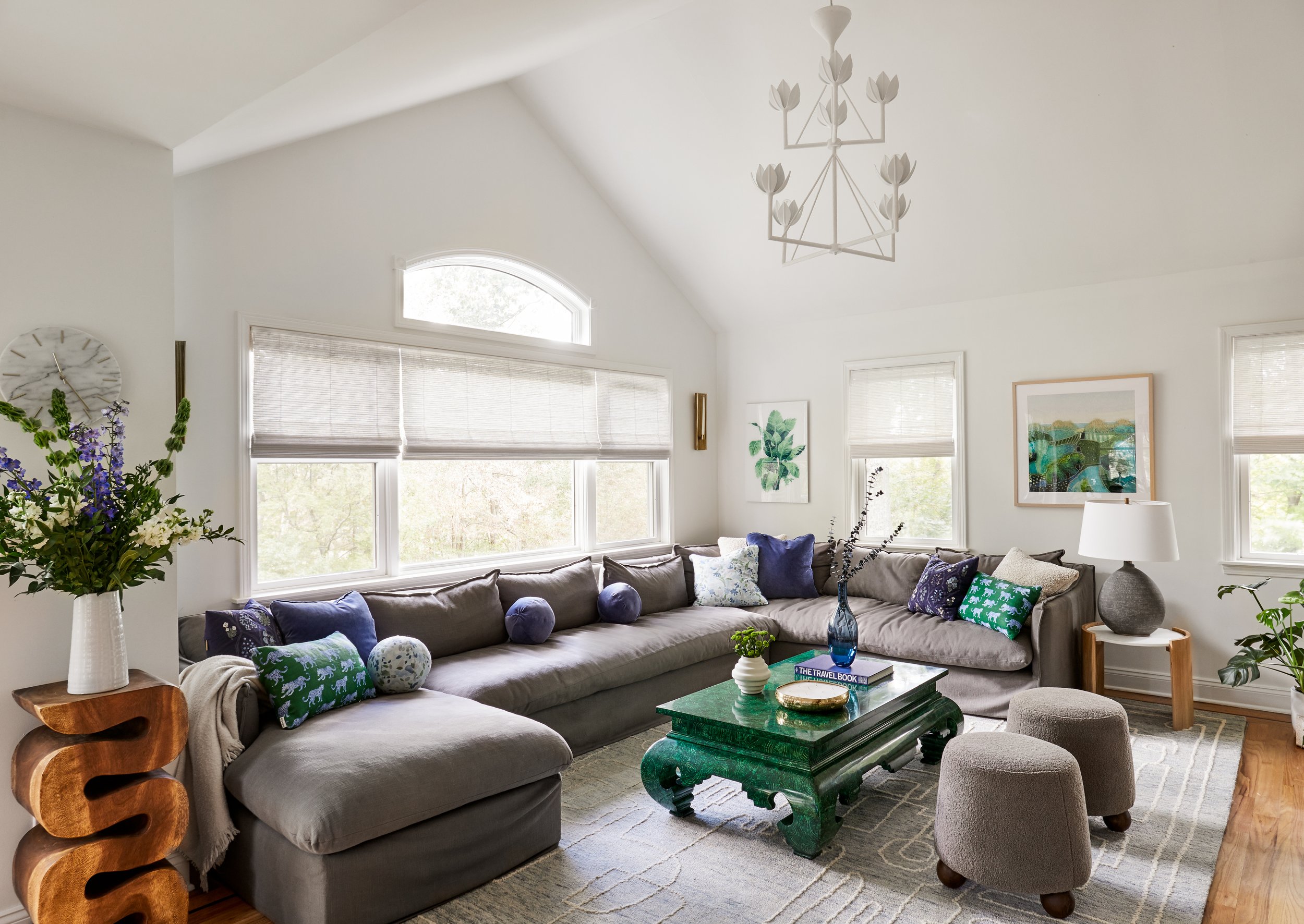 Living room with a large sectional sofa, colorful cushions, a green coffee table, and art on the walls, illuminated by multiple windows, with a chandelier hanging from the ceiling.