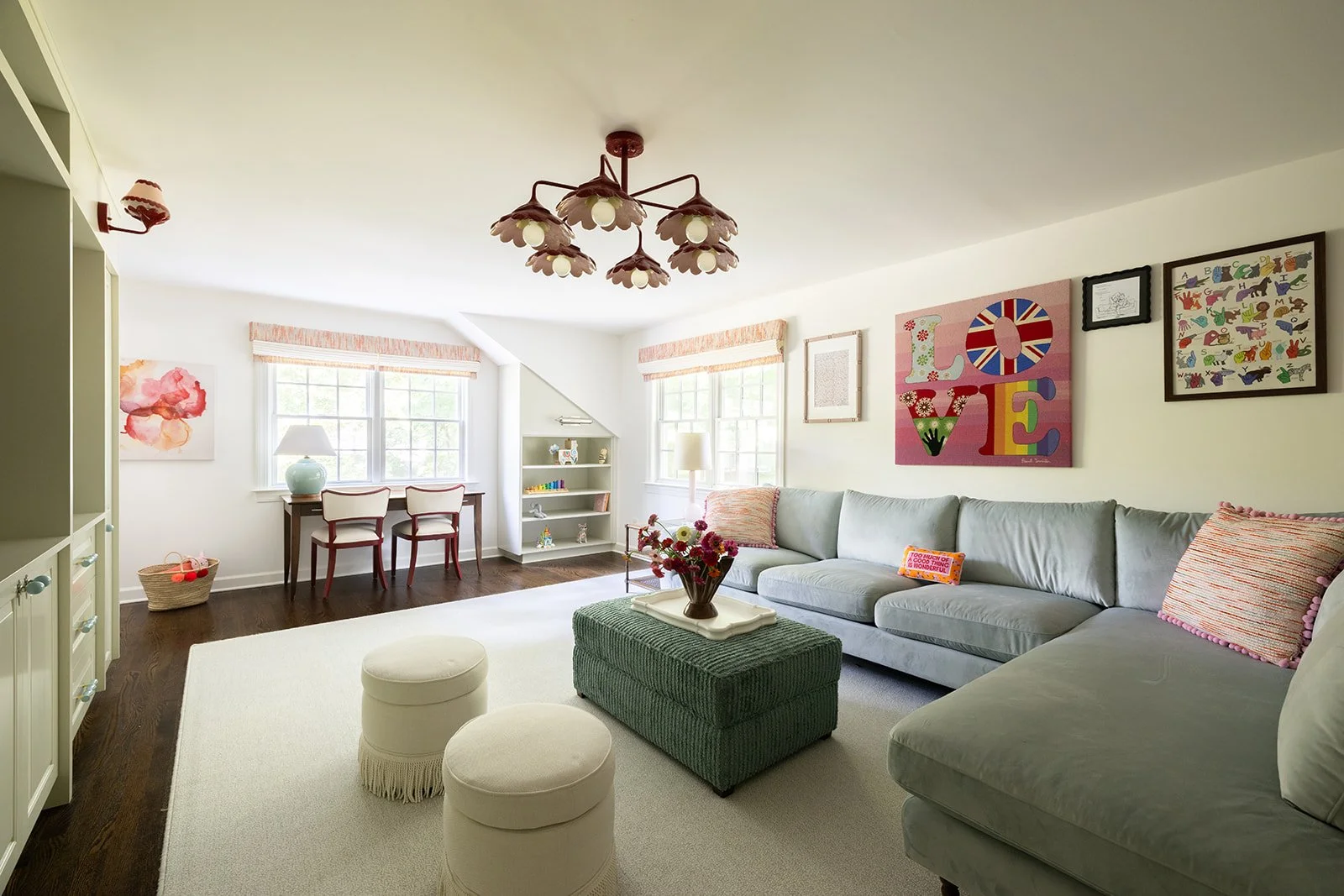 Bright living room with a large light blue sofa, colorful wall art, a dark wood floor, a beige area rug, and a small green ottoman with a tray and flowers. There are two white ottomans, a white bookshelf with decor, a small table and chairs by a wind