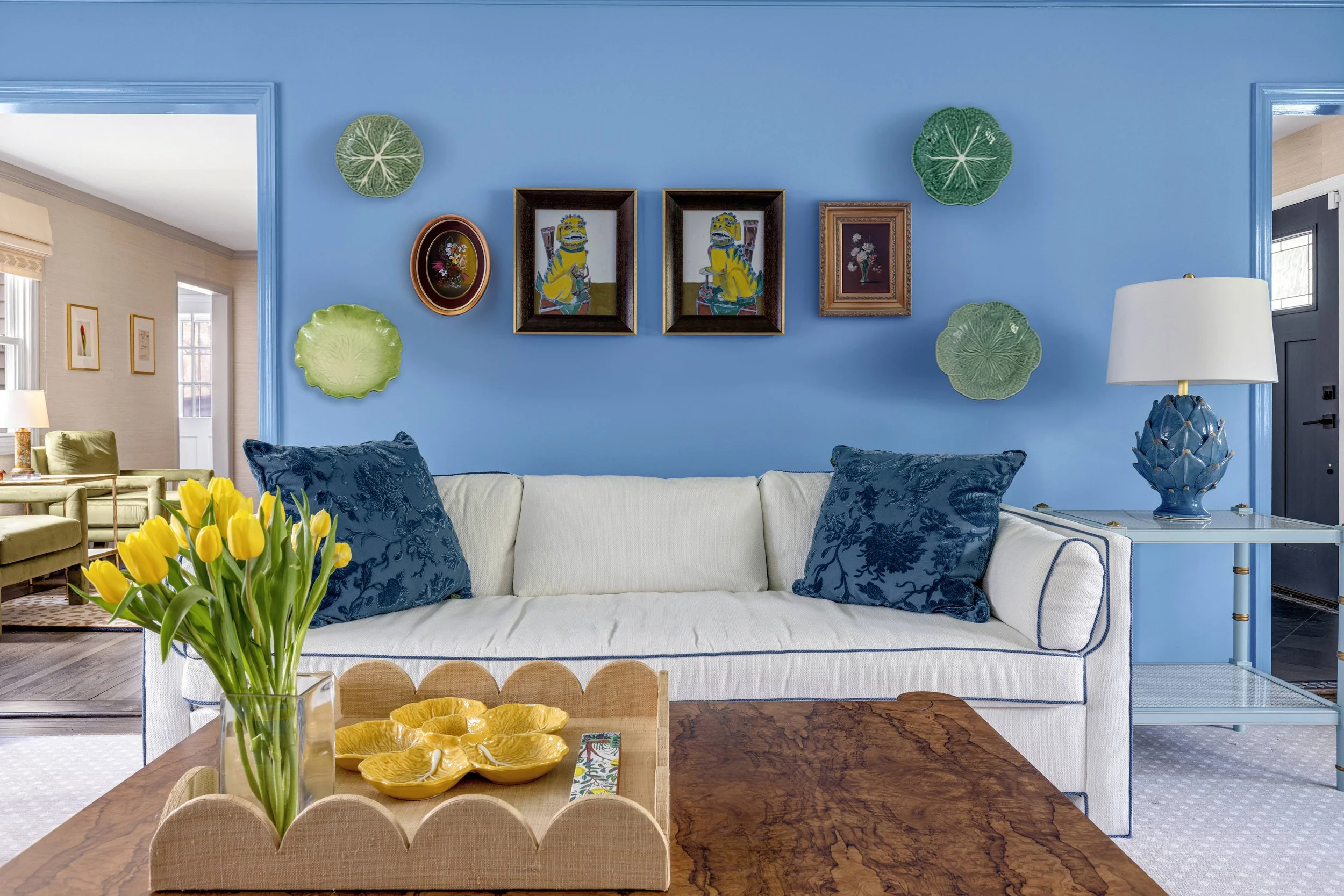 Living room with blue accent wall, framed artwork, green leaf wall decorations, white sofa with blue pillows, side table with lamp, wooden dining table with yellow tulips and decorative bowl.