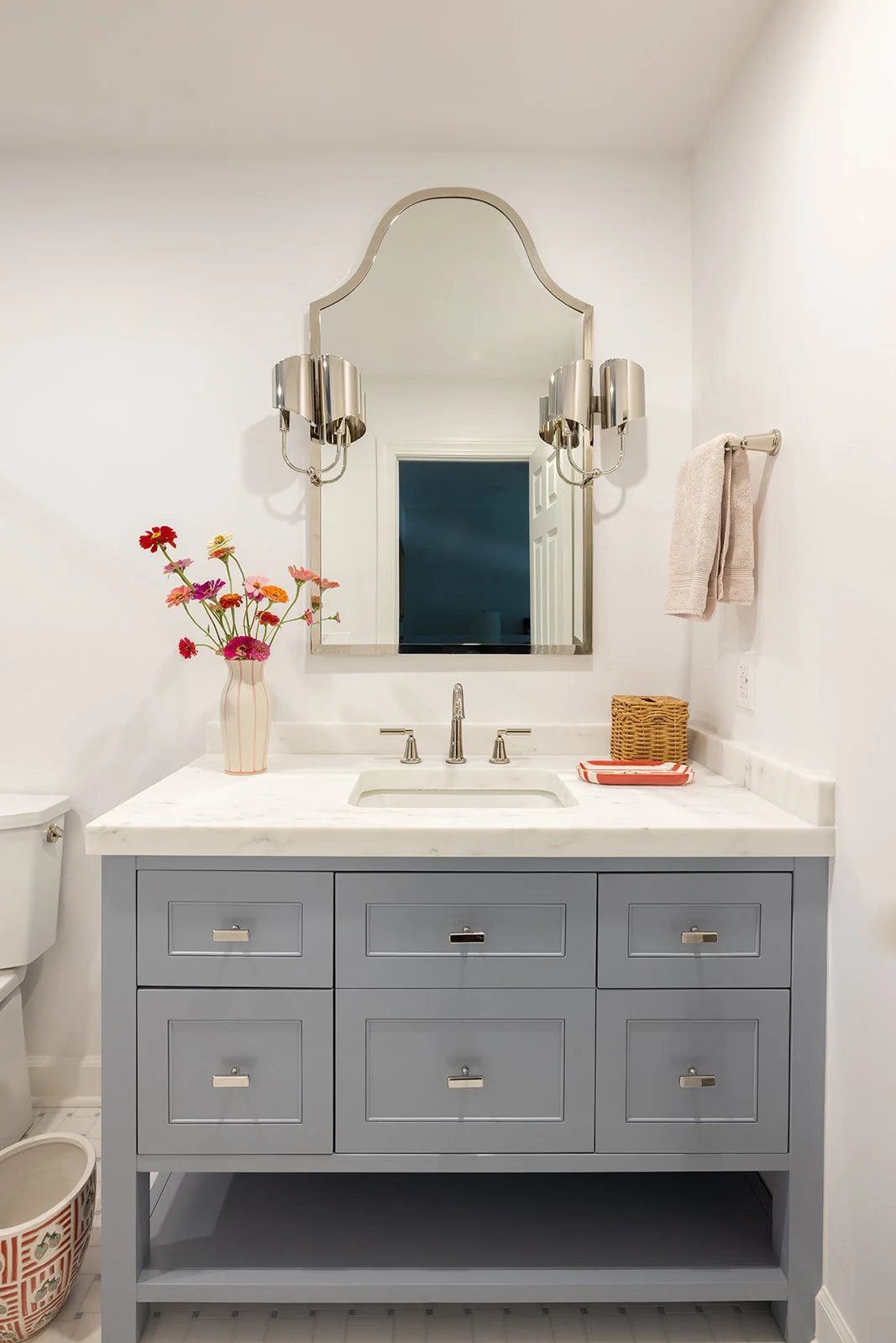 Bathroom vanity with a large mirror, silver light fixtures, a vase with pink, orange, and purple flowers, towels, and a wicker basket on a white marble countertop.