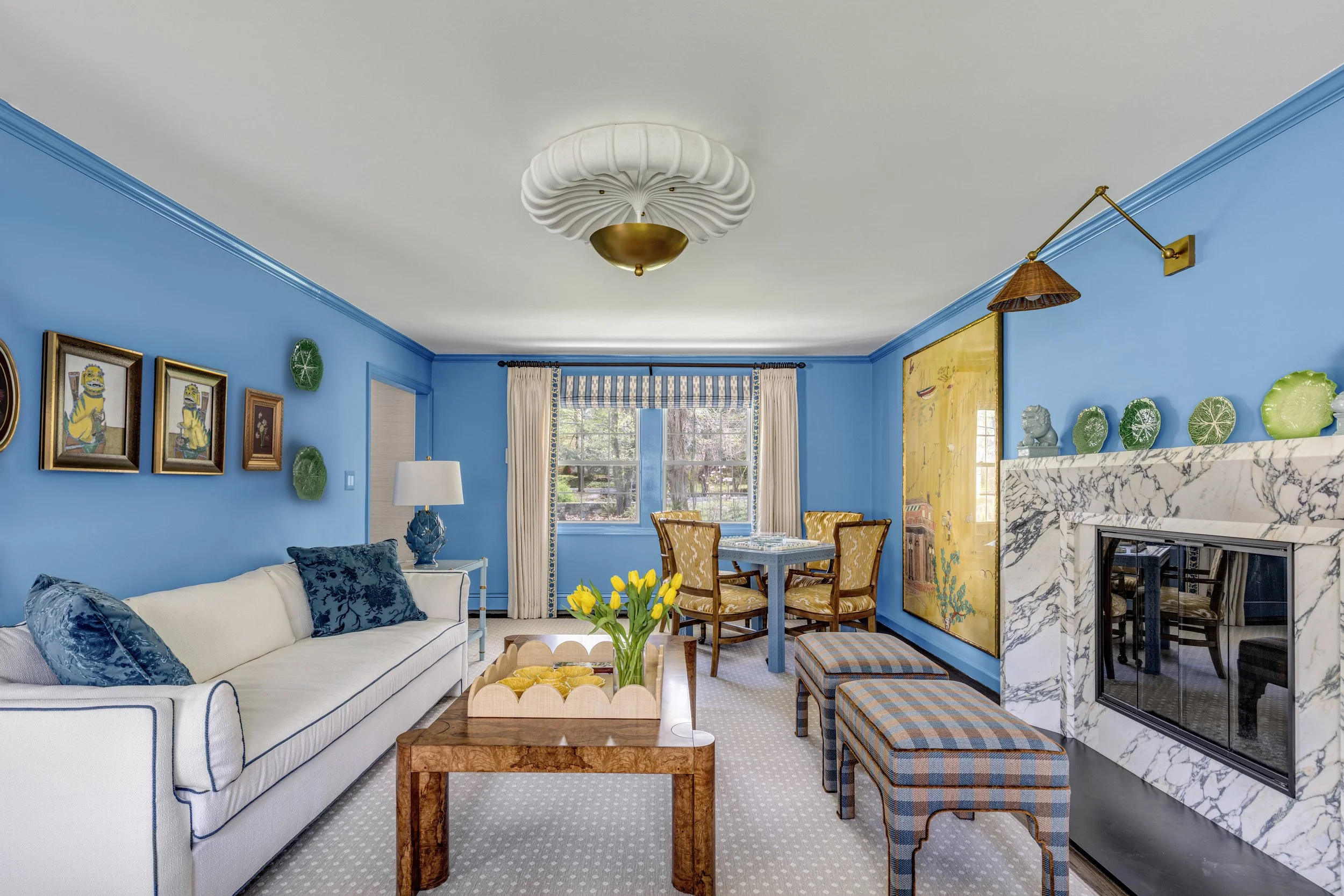 Living room with blue walls, white sofa with blue pillows, marble fireplace, yellow armchairs, and a dining table with chairs. Decor includes framed artwork, green wall ornaments, and a large window with white curtains.