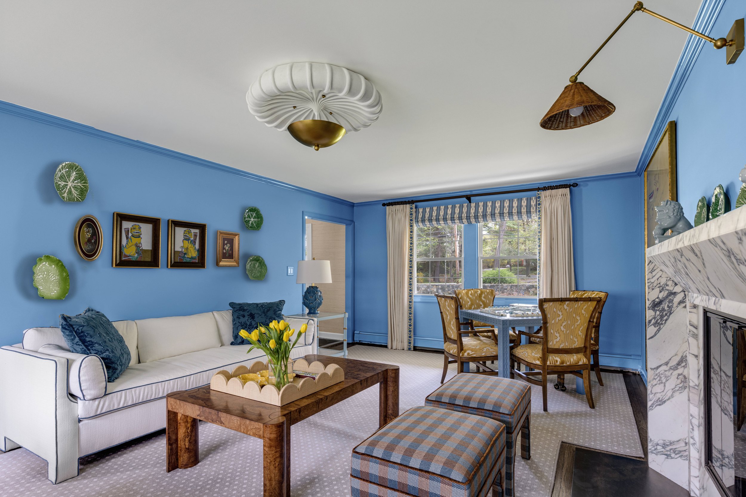 Living room with blue walls, white couch with blue pillows, wooden coffee table with yellow tulips, patterned window curtains, and wall art featuring a character resembling a Minion. Ceiling has decorative lighting fixture, and there is a wall-mounte