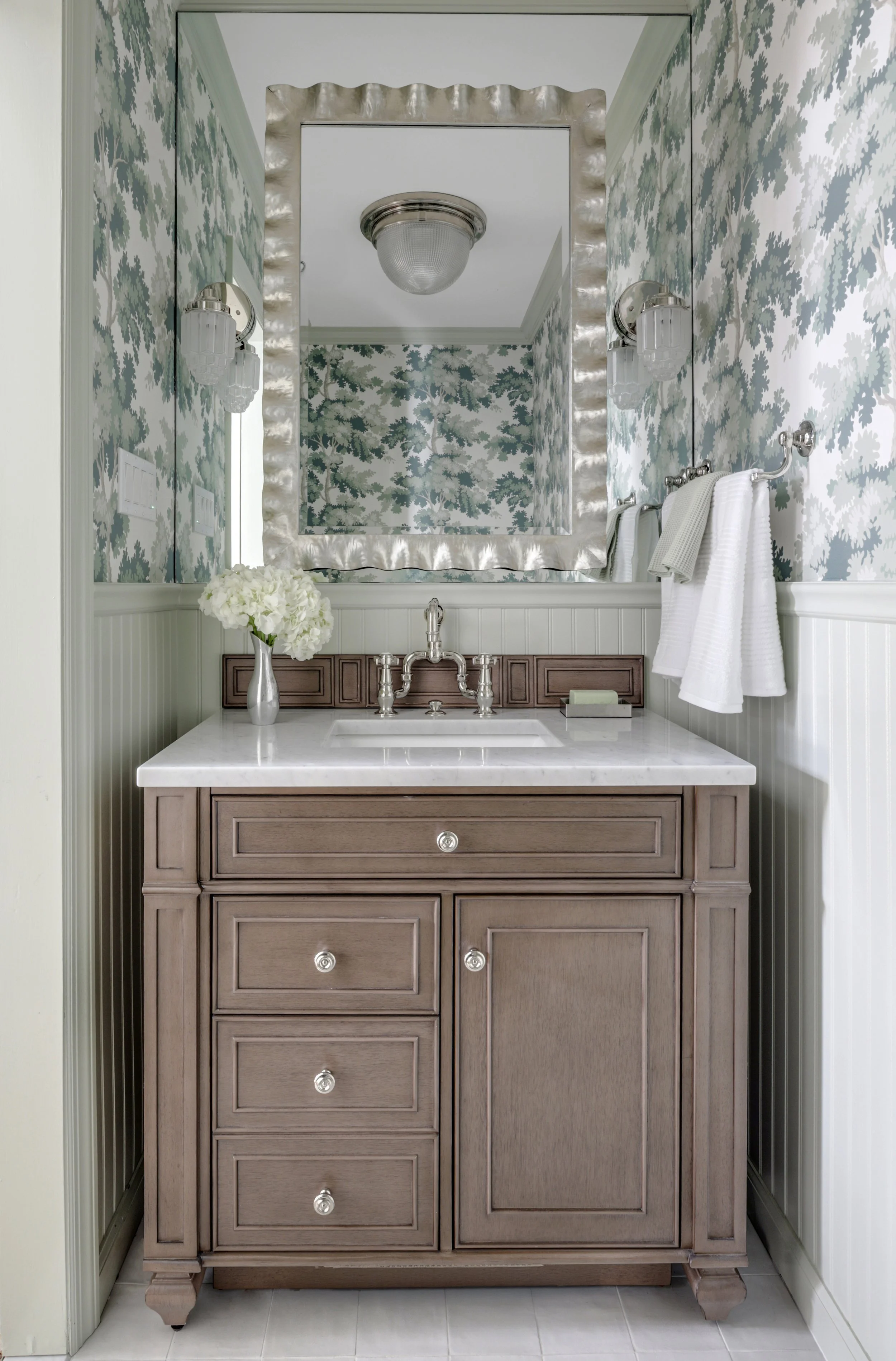 A bathroom vanity with a light wood finish, a white marble countertop, a sink, vintage-style silver faucet, and a mirror framed in fabric with a metallic sheen. There is a small floral arrangement and soap on the countertop. The wall has wallpaper with a blue and white floral pattern, and there are two wall-mounted lights.