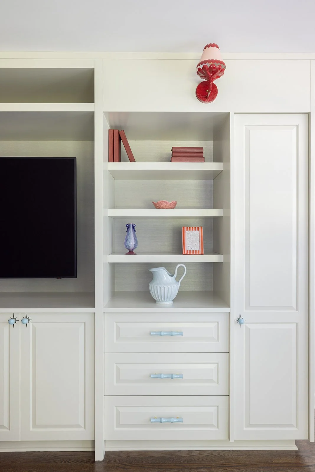 A white built-in bookshelf with decorative items, a small television on the left, and a closed cabinet door on the right. The shelves contain books, a pink bowl, a blue vase, a framed picture, a white pitcher, and a pink and red wall-mounted lamp on 