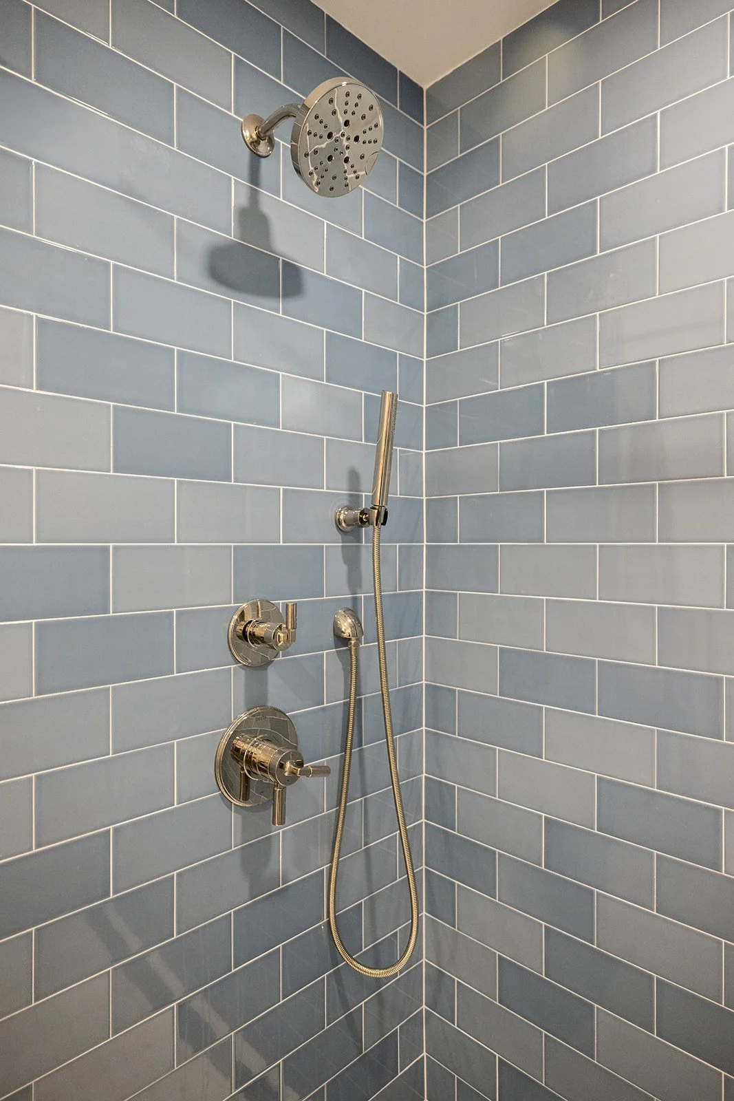 A shower corner with blue subway tiles, a large round showerhead, a handheld shower spray, and control knobs in polished metal.