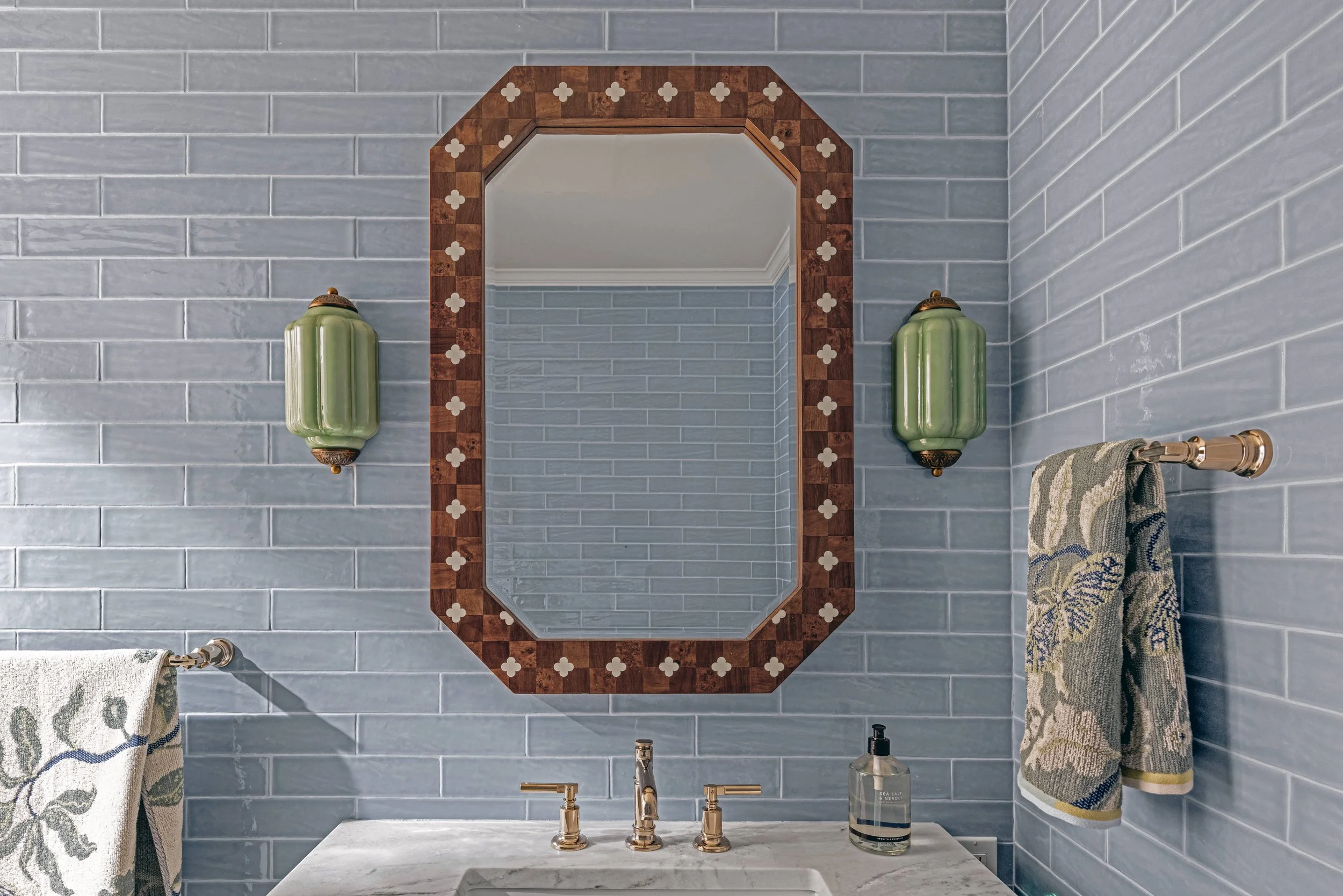 A bathroom with blue subway tiles, a framed mirror, two green wall-mounted light fixtures, a towel rack with a patterned towel, a marble countertop with a gold faucet, a soap dispenser, and a rolled-up towel.