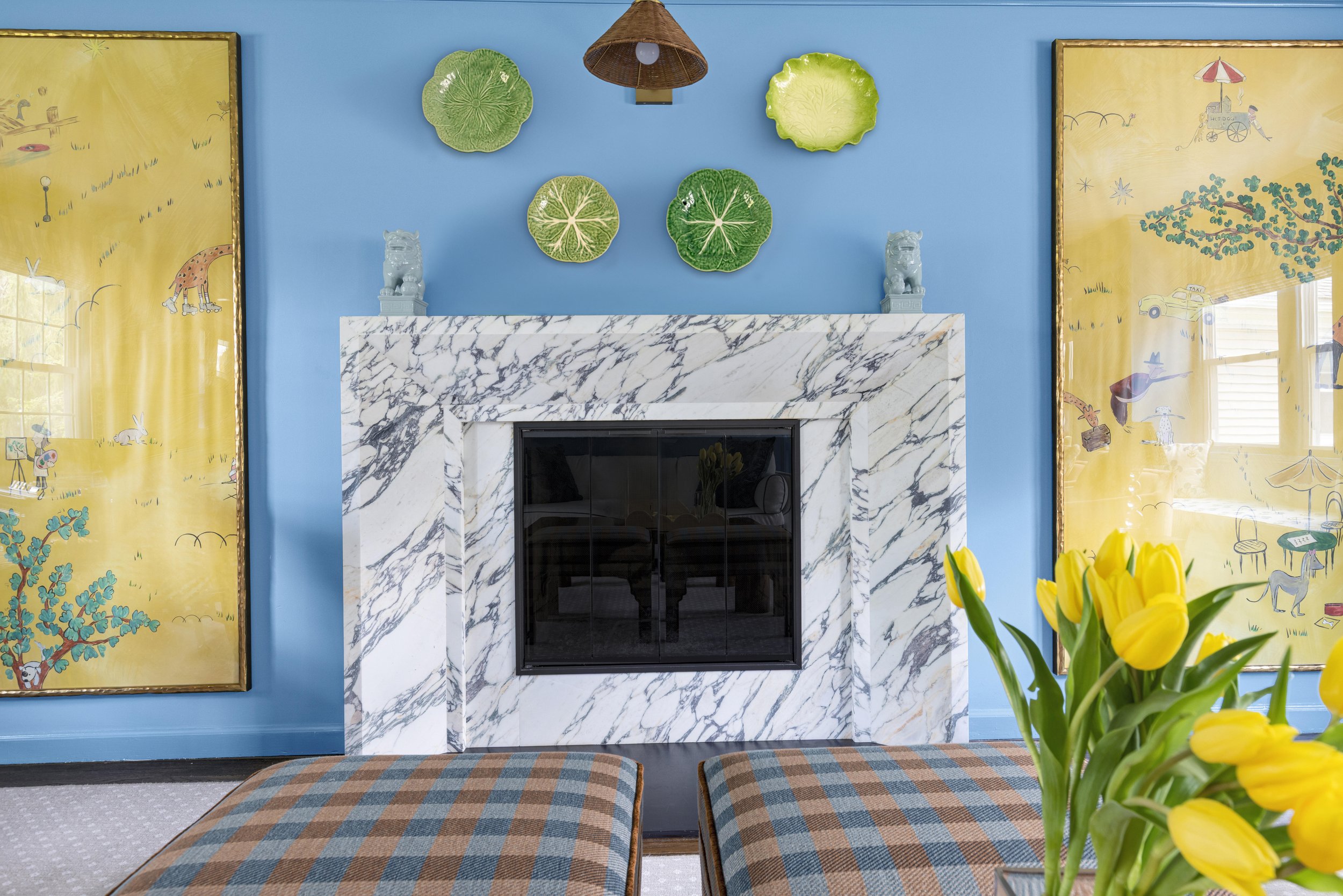 A living room fireplace with a white and black marble mantel, decorated with two small statues of lions. Above the mantel, the wall is painted blue and displays four decorative green and yellow leaf-shaped plates. Flanking the fireplace are two yello