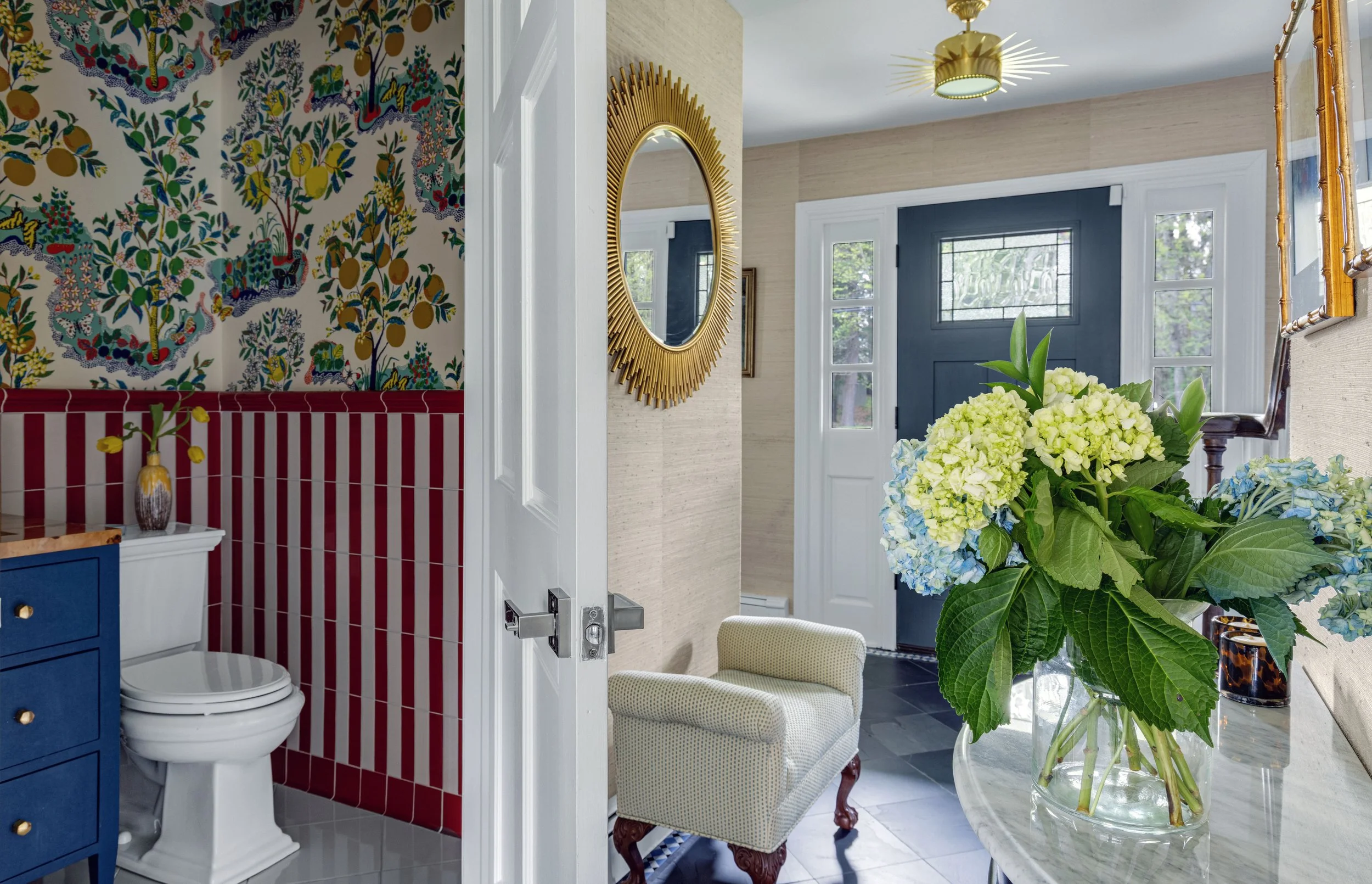 Interior view of a room with a large bouquet of white and green hydrangeas in a clear glass vase on a marble table, a mirror with a gold sunburst frame on the wall, a gold ceiling light, a blue door with glass panes, a small beige armchair with a cur