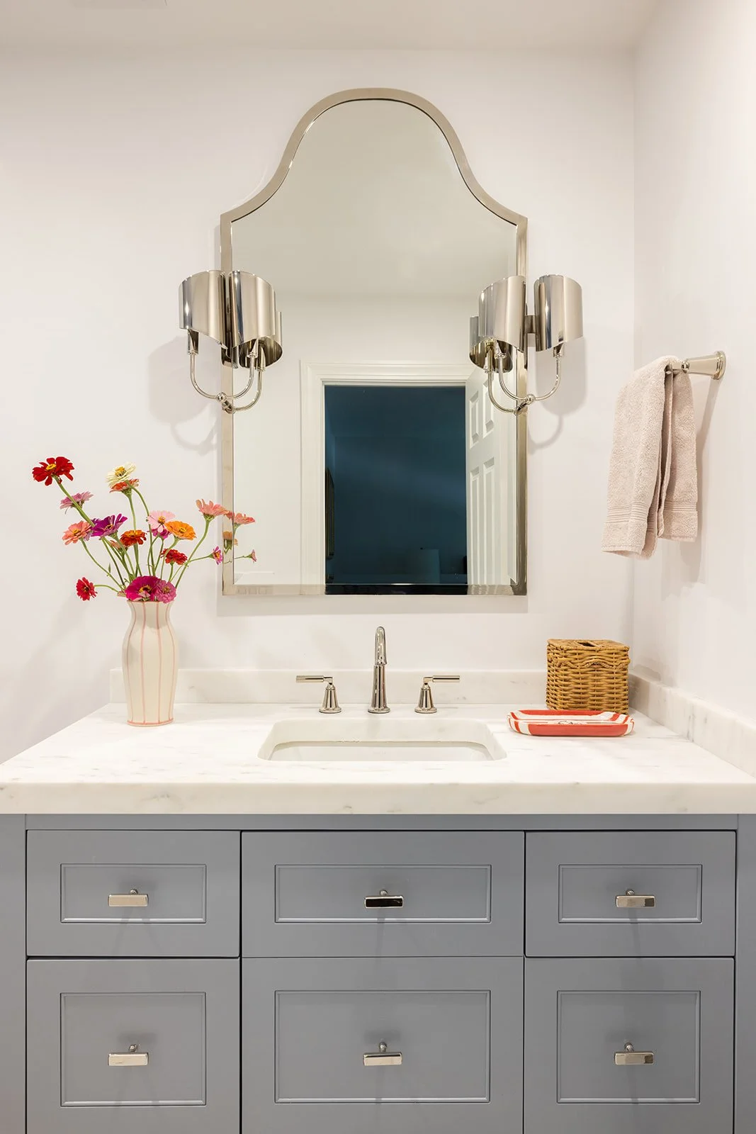 Bathroom vanity with gray drawers, white marble countertop, mirror, vase with colorful flowers, towel, and wicker basket.