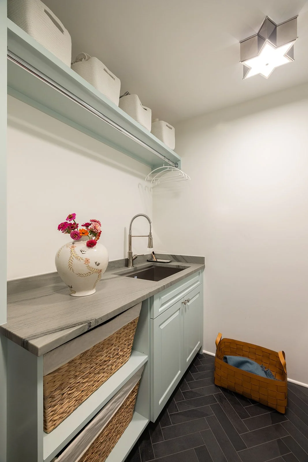 Small laundry or utility room with pale green cabinetry, a marble countertop, a small sink with a modern faucet, a decorative vase with pink flowers, white storage baskets on a shelf above, and a yellow woven basket on the floor. There is a ceiling l