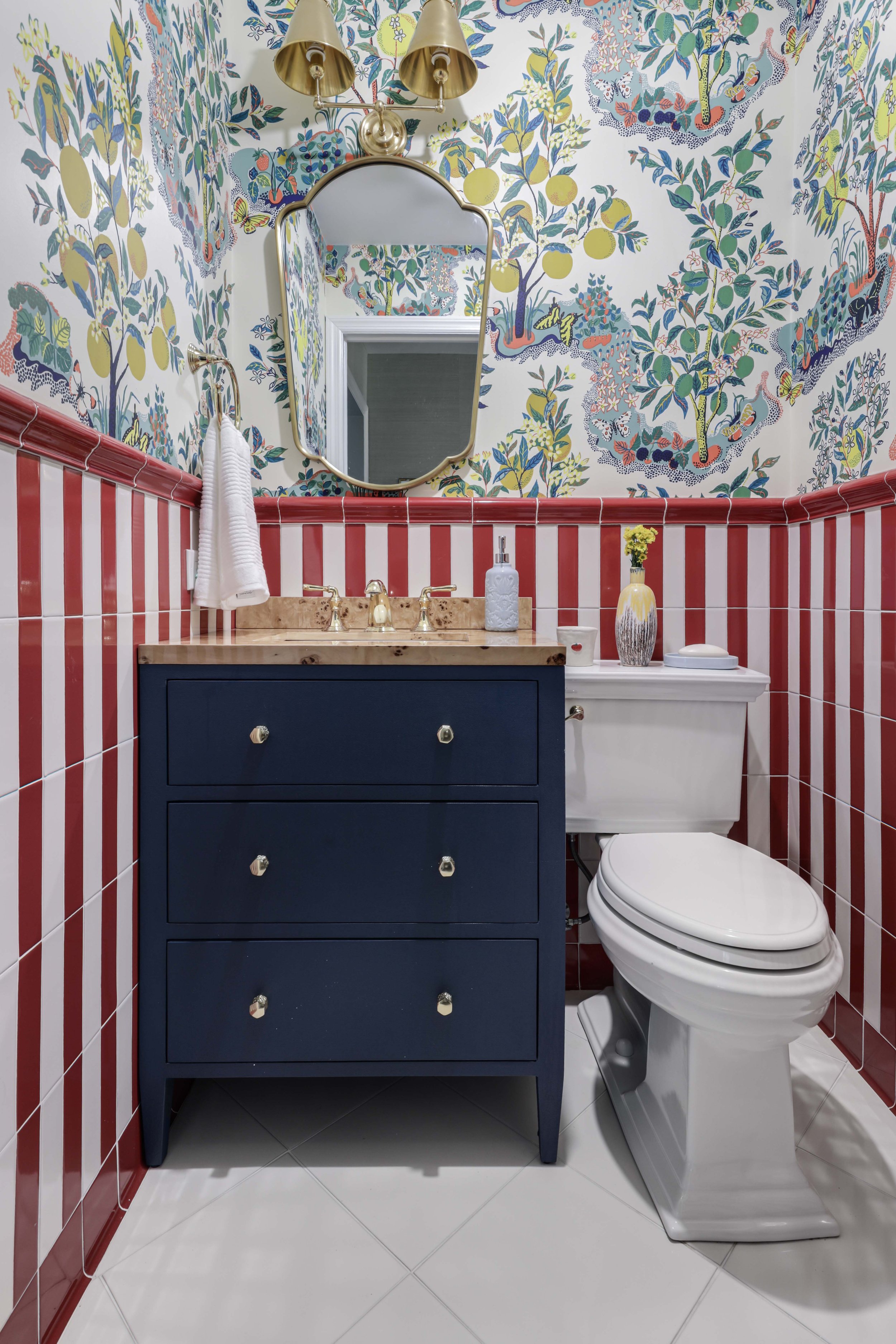 A colorful bathroom with floral wallpaper, red and white tiled walls, a dark blue vanity with a wooden countertop, a mirror, a gold wall-mounted light fixture, and a white toilet. There are decorative items like a vase with yellow flowers and a soap 