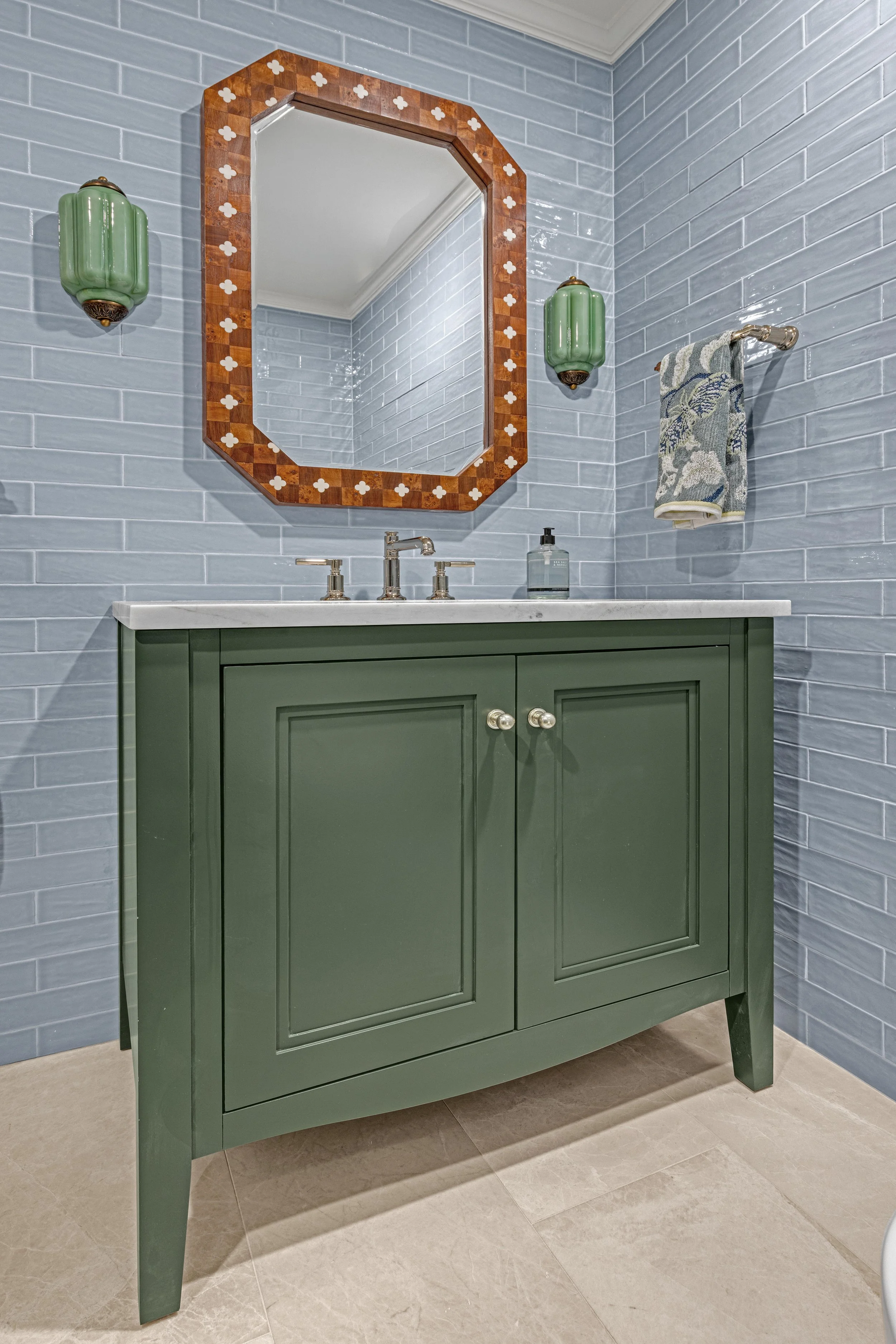 A green bathroom vanity with a white marble countertop, silver faucet, and a soap dispenser. Above it is a wooden framed mirror with white cross inlays, flanked by two green wall sconces. There is a towel hanging on a silver bar and blue tiled walls.