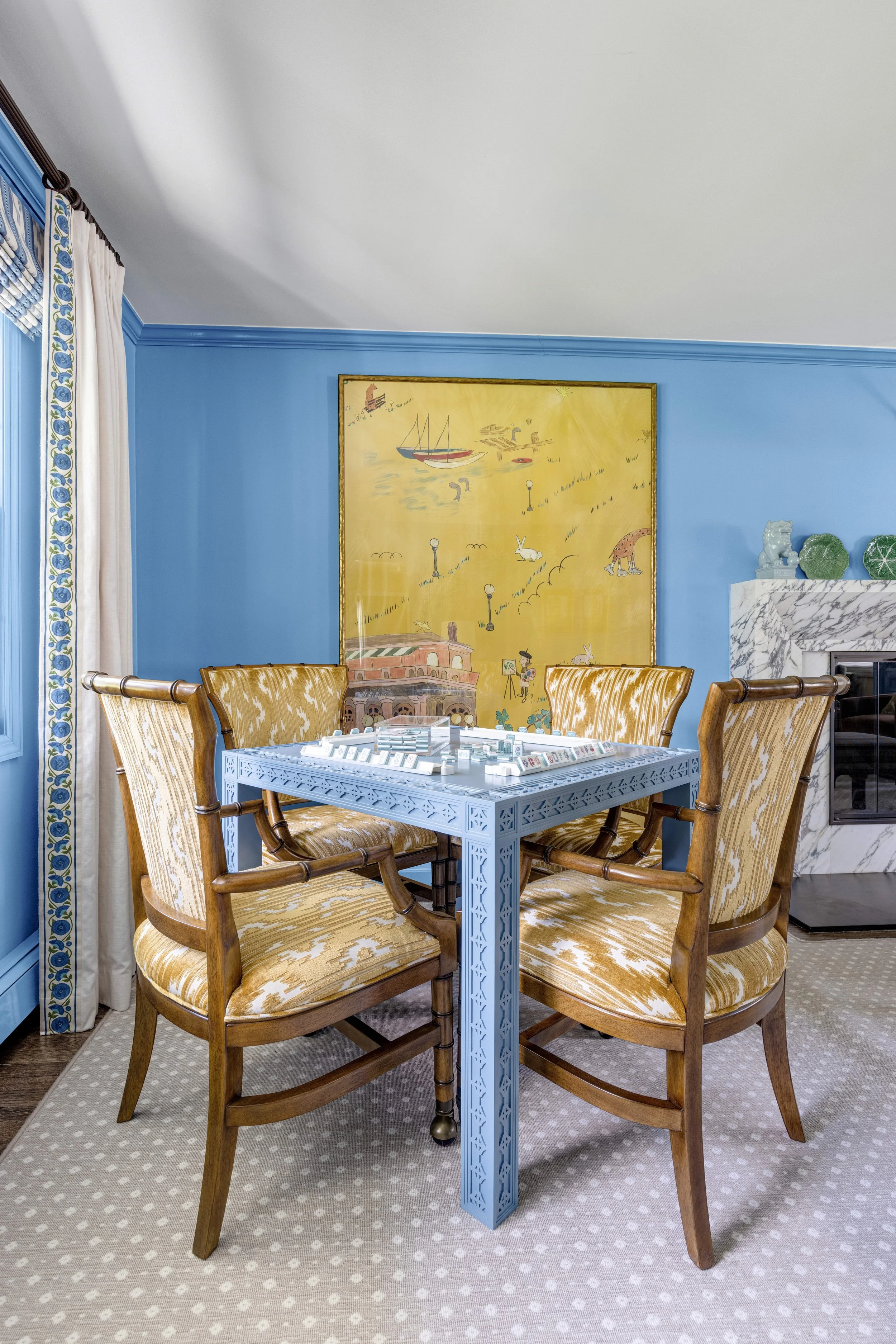 Dining room with a round table, six wooden upholstered chairs, a yellow decorative painting on the blue wall, and a marble fireplace with decorative objects.