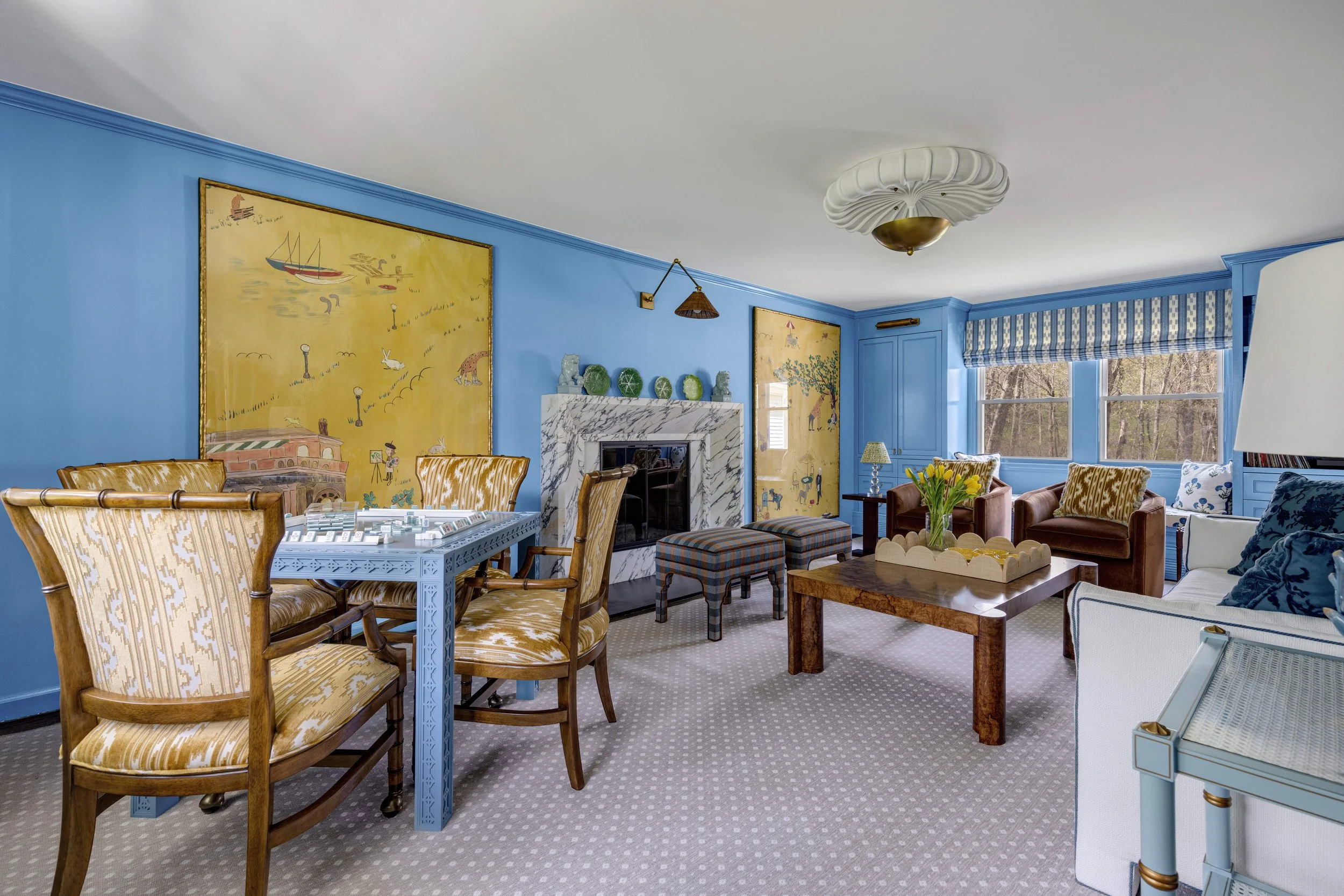 Living room with blue walls, decorated with two large paintings, a marble fireplace, multiple chairs, sofas, a coffee table with a vase of yellow tulips, a ceiling light fixture, and large windows with blue striped curtains, overlooking trees outside