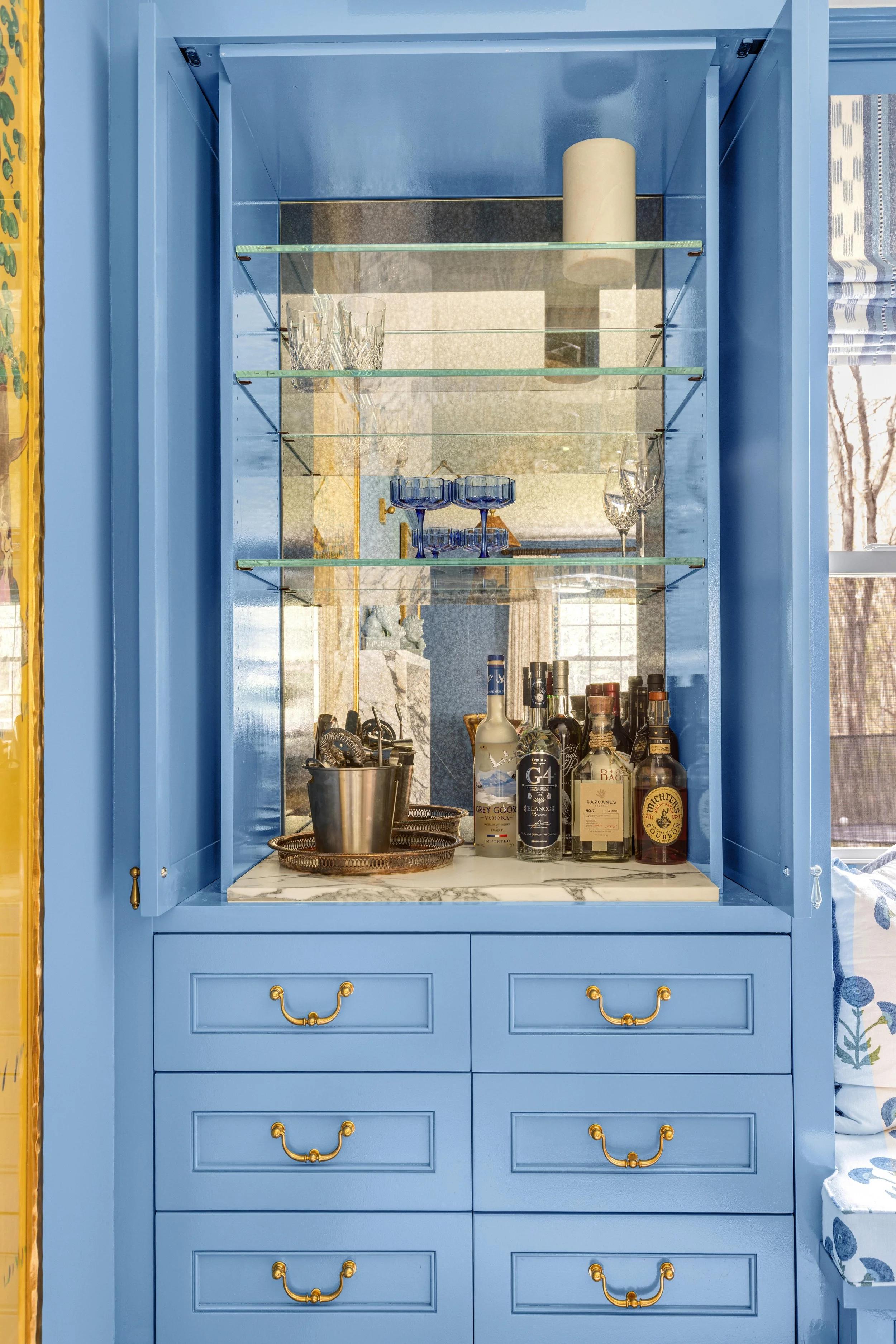A bright blue bar cabinet with glass shelves, displaying glassware and liquor bottles, topped with a marble slab.