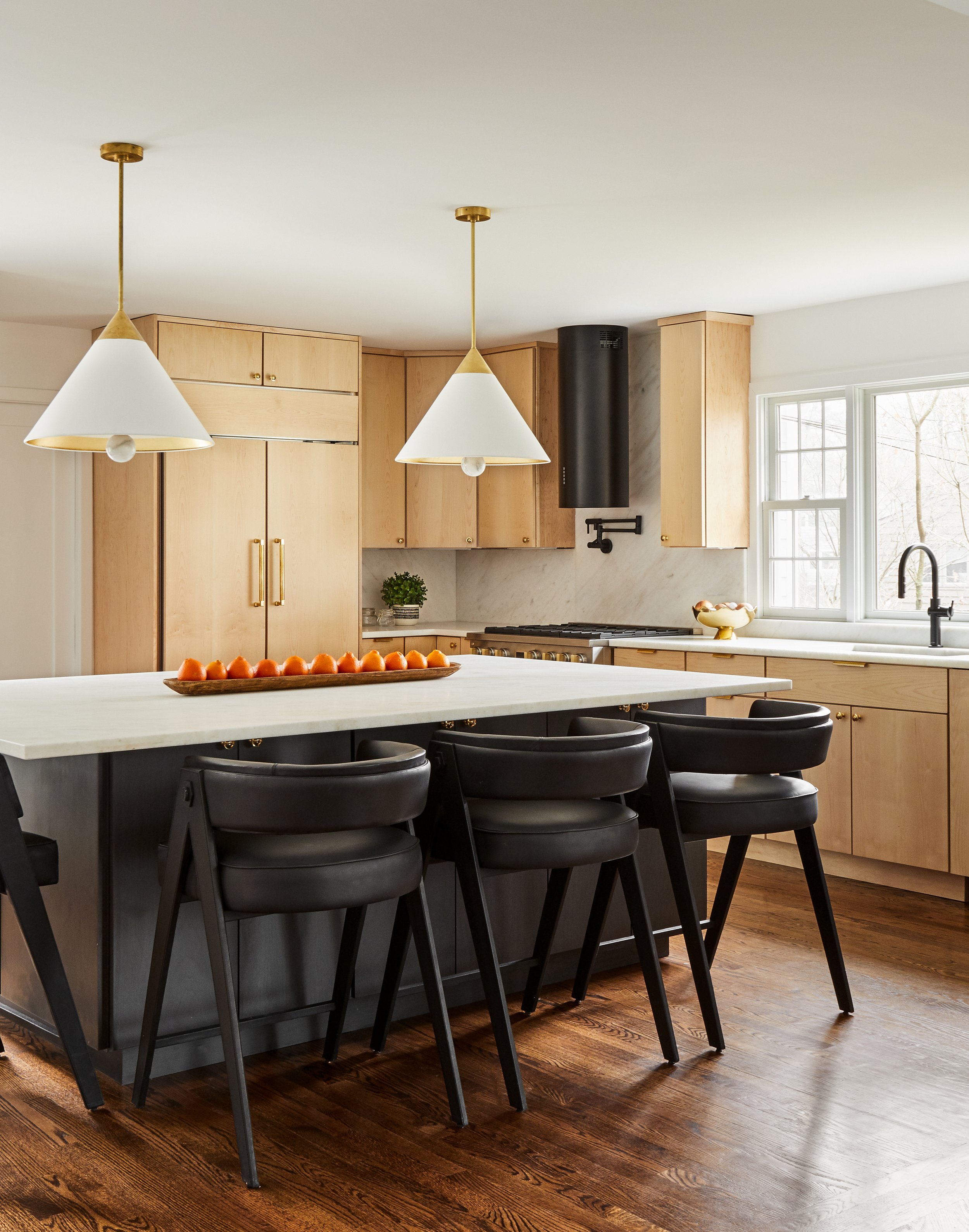 Modern kitchen with light wood cabinets, black island with black chairs, marble countertops, two white pendant lights, a window, and a black stove hood.