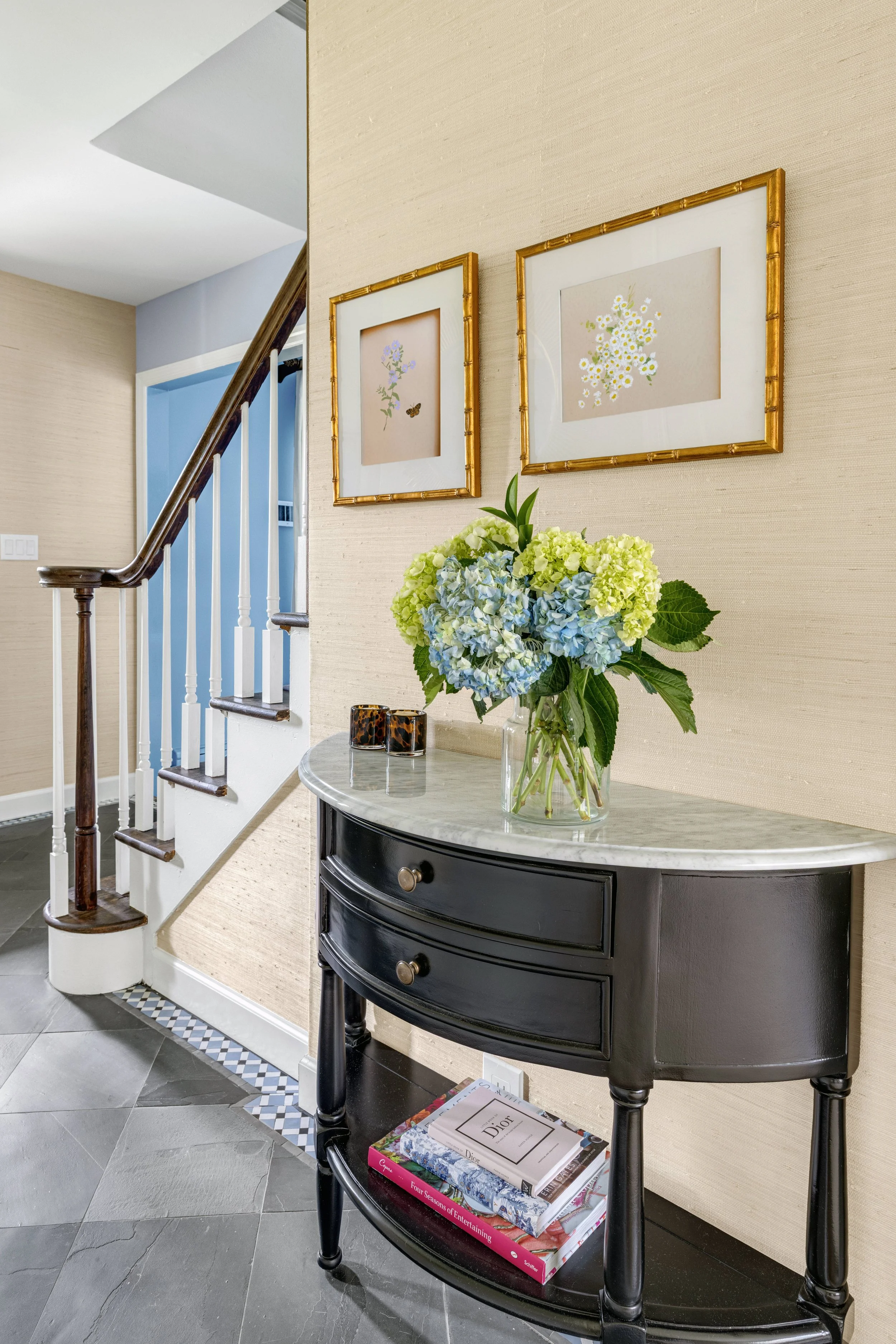 A black, curved console table with drawers and gold knobs, placed against a beige wall with framed botanical artwork. A large bouquet of white and blue hydrangeas sits on top, along with several books on the bottom shelf. The corner of a staircase wi