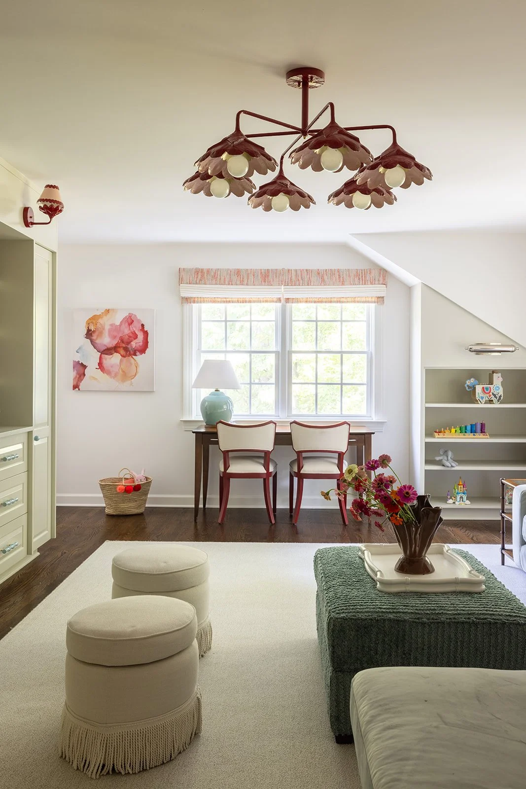 A cozy, well-lit living room with a large window, a chandelier with floral-shaped light fixtures, a white carpet, and various furniture and decorative items including a green ottoman, a tray with a flower vase, a desk with two chairs, and a bookshelf