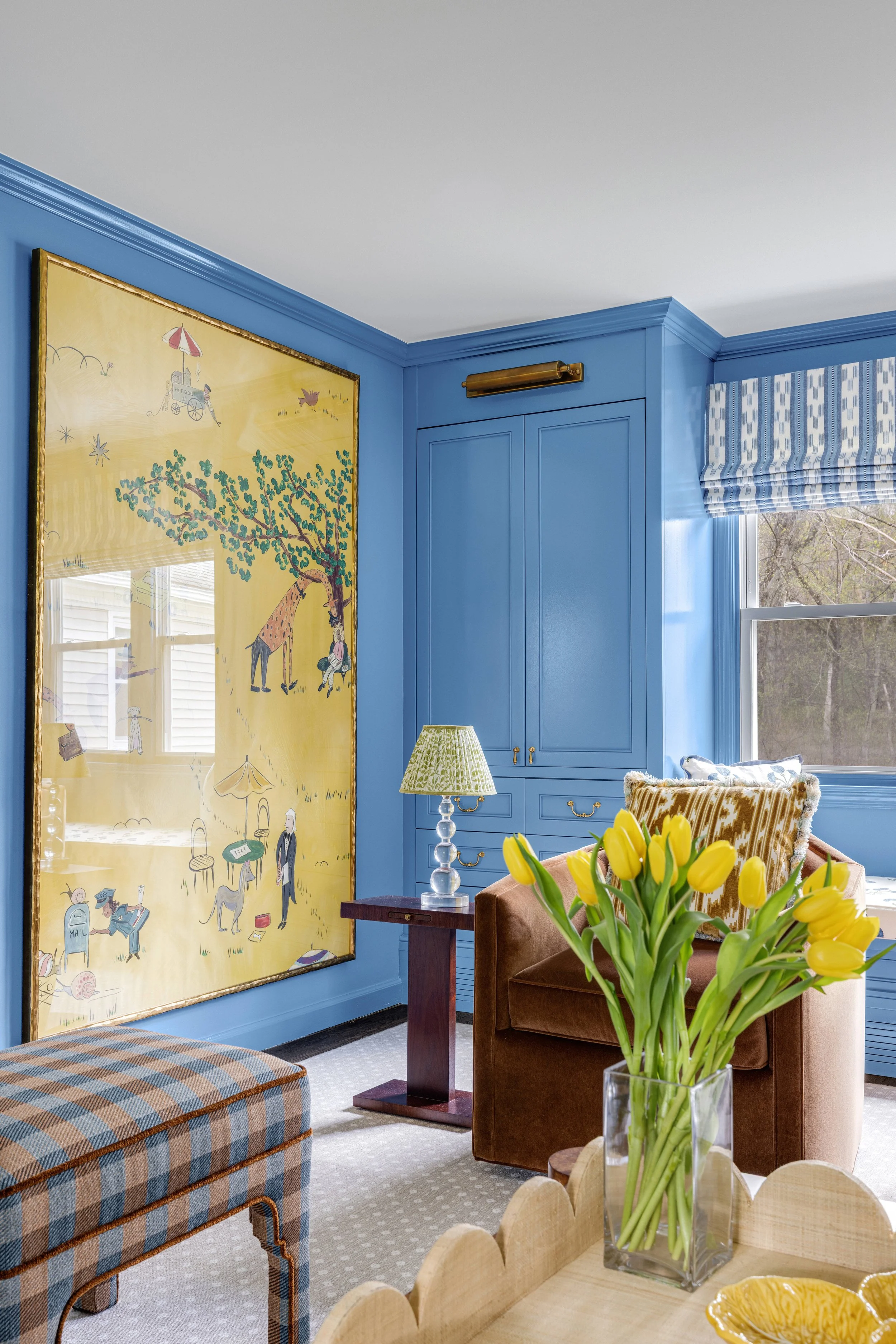 A cozy living room with a blue wall, a large framed artwork, a window with blue and white striped shades, and a bouquet of yellow tulips on a table. There is a brown armchair, a table lamp with a green patterned shade, and a plaid upholstered bench.