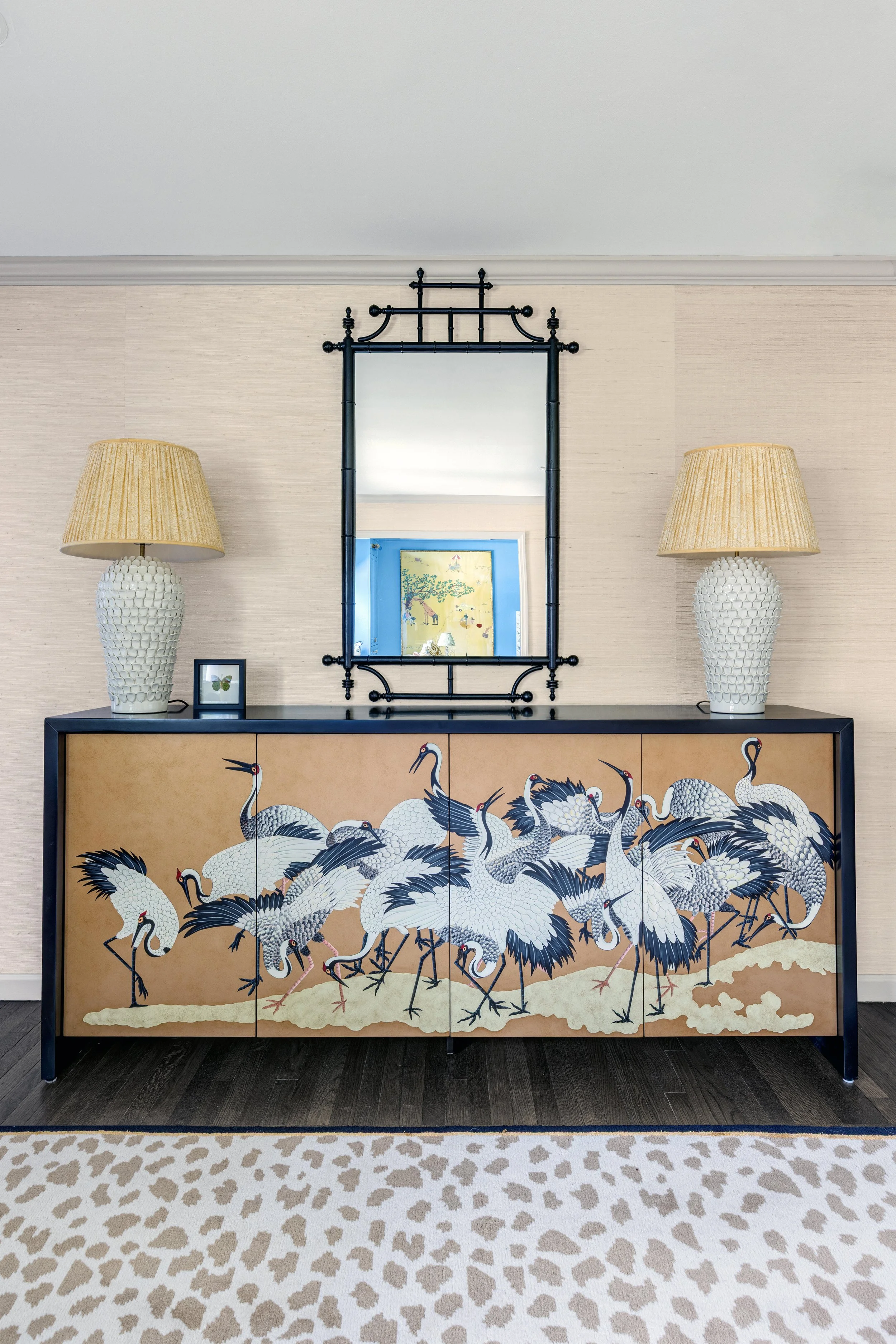 A decorative cabinet with a crane design and a large mirror on top, flanked by two table lamps with beige shades, set against a plain wall with a leopard print rug on the floor.