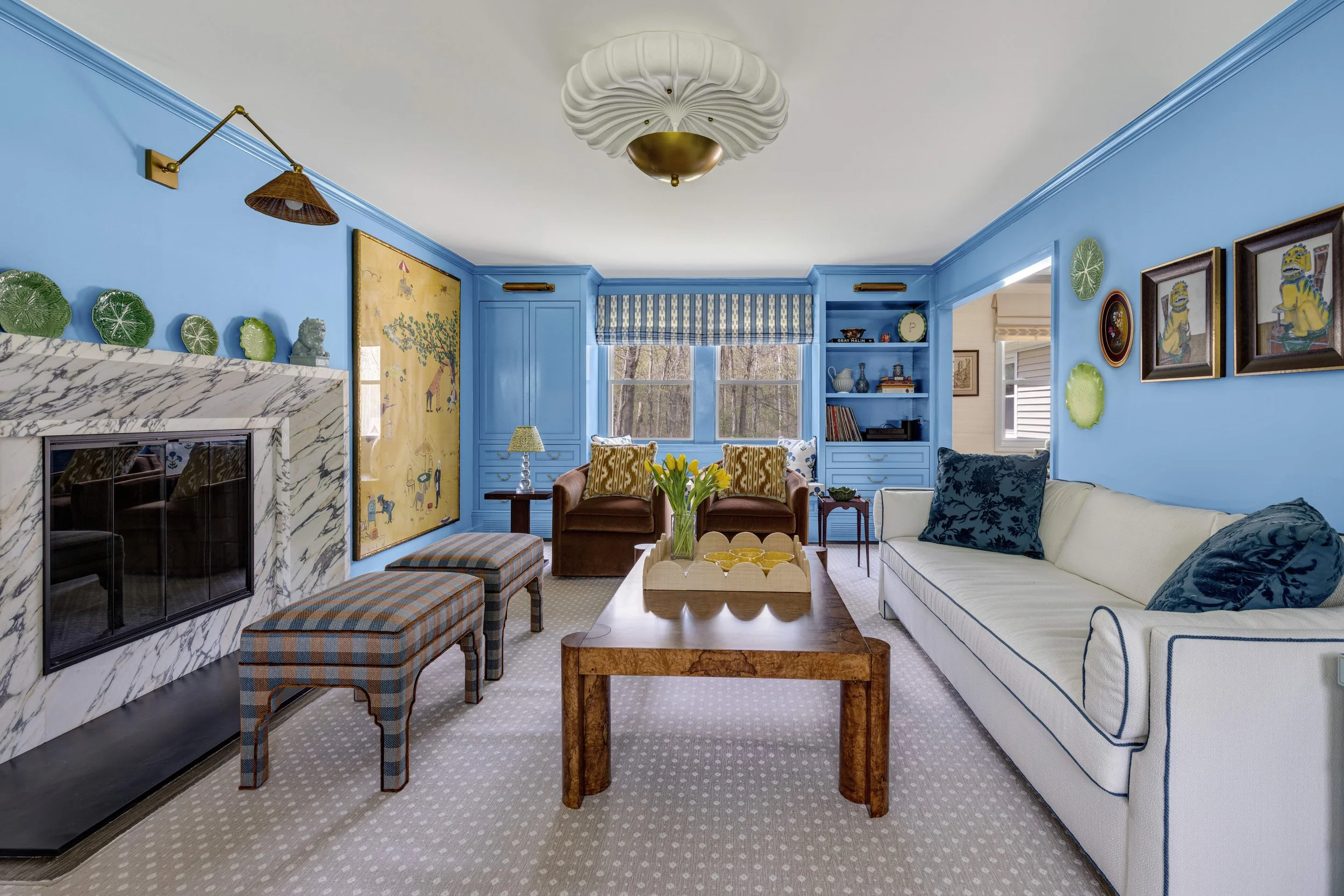Living room with blue walls, a white ceiling, and multiple chairs and sofas. A marble fireplace is on the left, with decorative plates and art pieces above it. A window with striped curtains and built-in shelves filled with books and decorative items