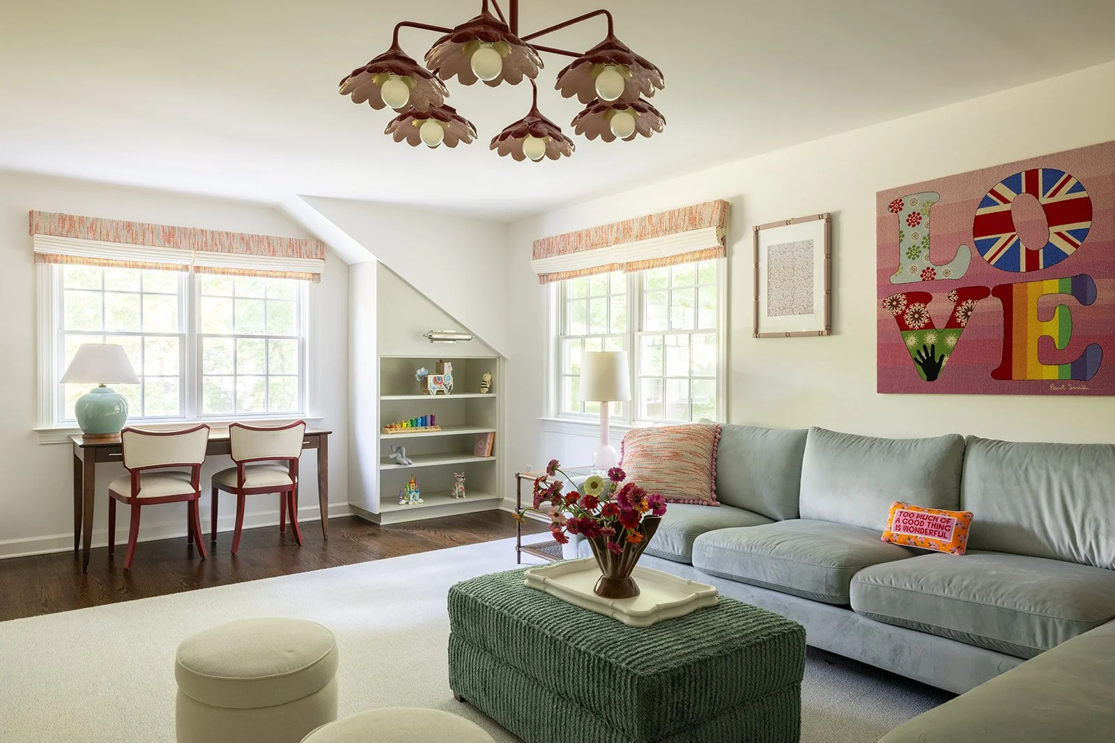 Bright living room with large windows, pastel sofa, floral artwork, white bookshelf with toys, and a table with a flower arrangement.