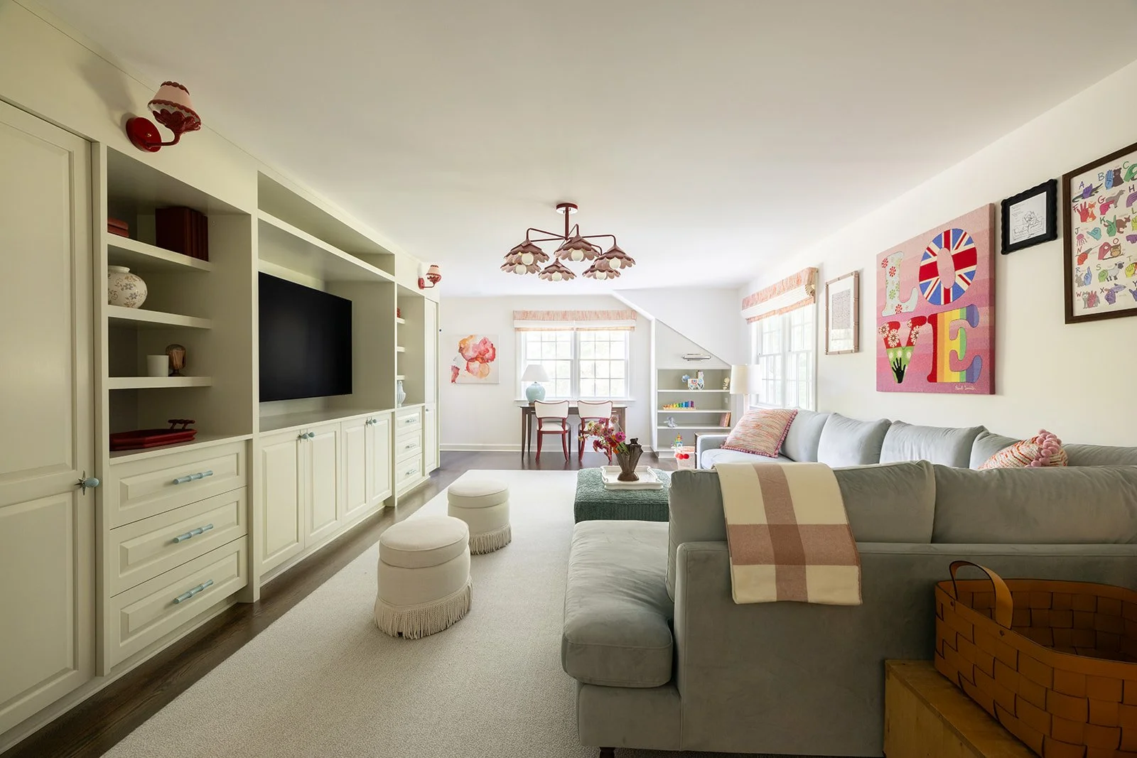 Bright living room with white built-in shelves, a gray sectional sofa with pastel cushions, colorful artwork on the walls, and a window with pink valances, creating a cozy and cheerful atmosphere.