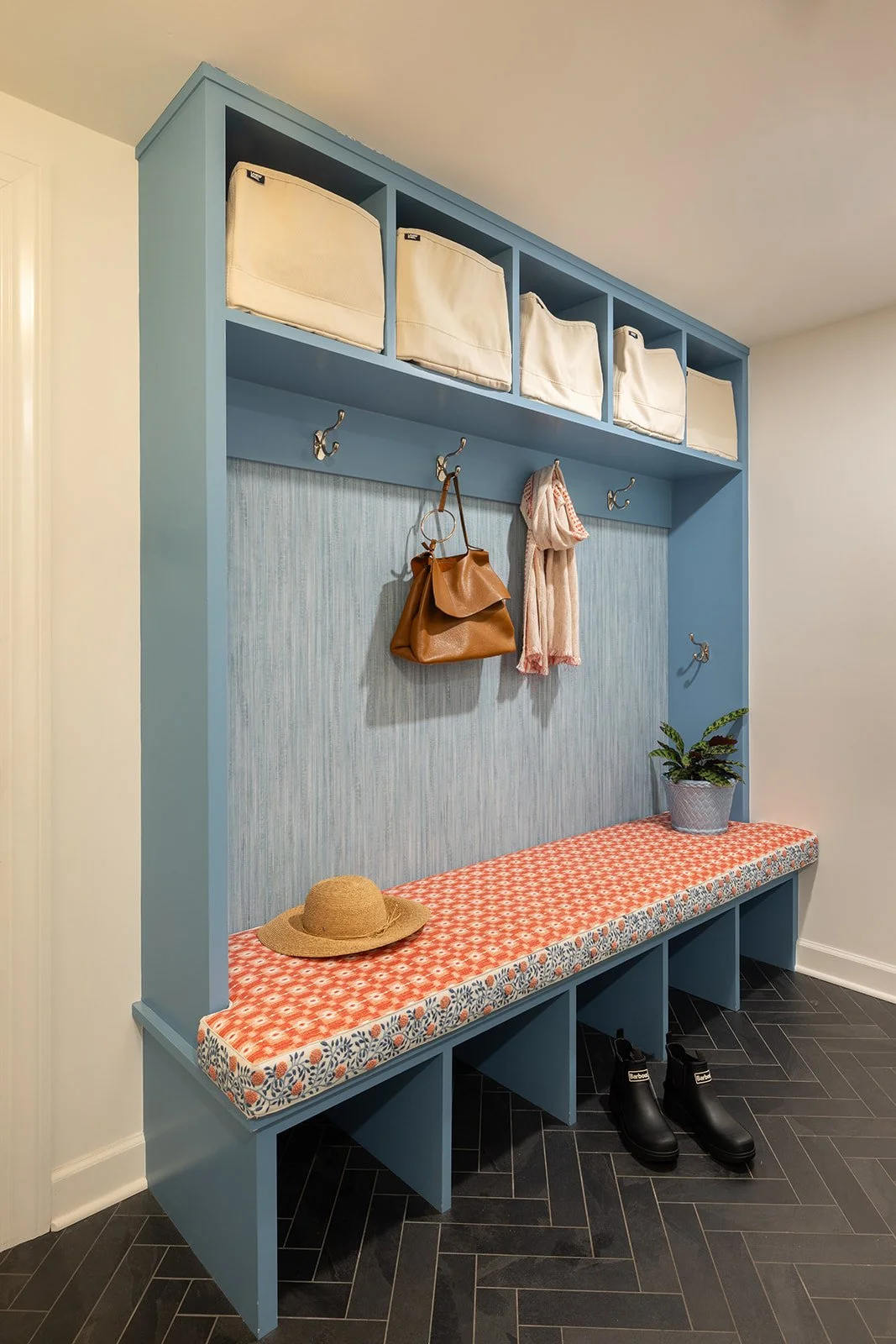 A blue entryway hall tree with a cushioned bench, a plant, a hat, and a pair of boots underneath. The back has hooks with a purse and scarf hanging from them, and a shelf with storage baskets above.