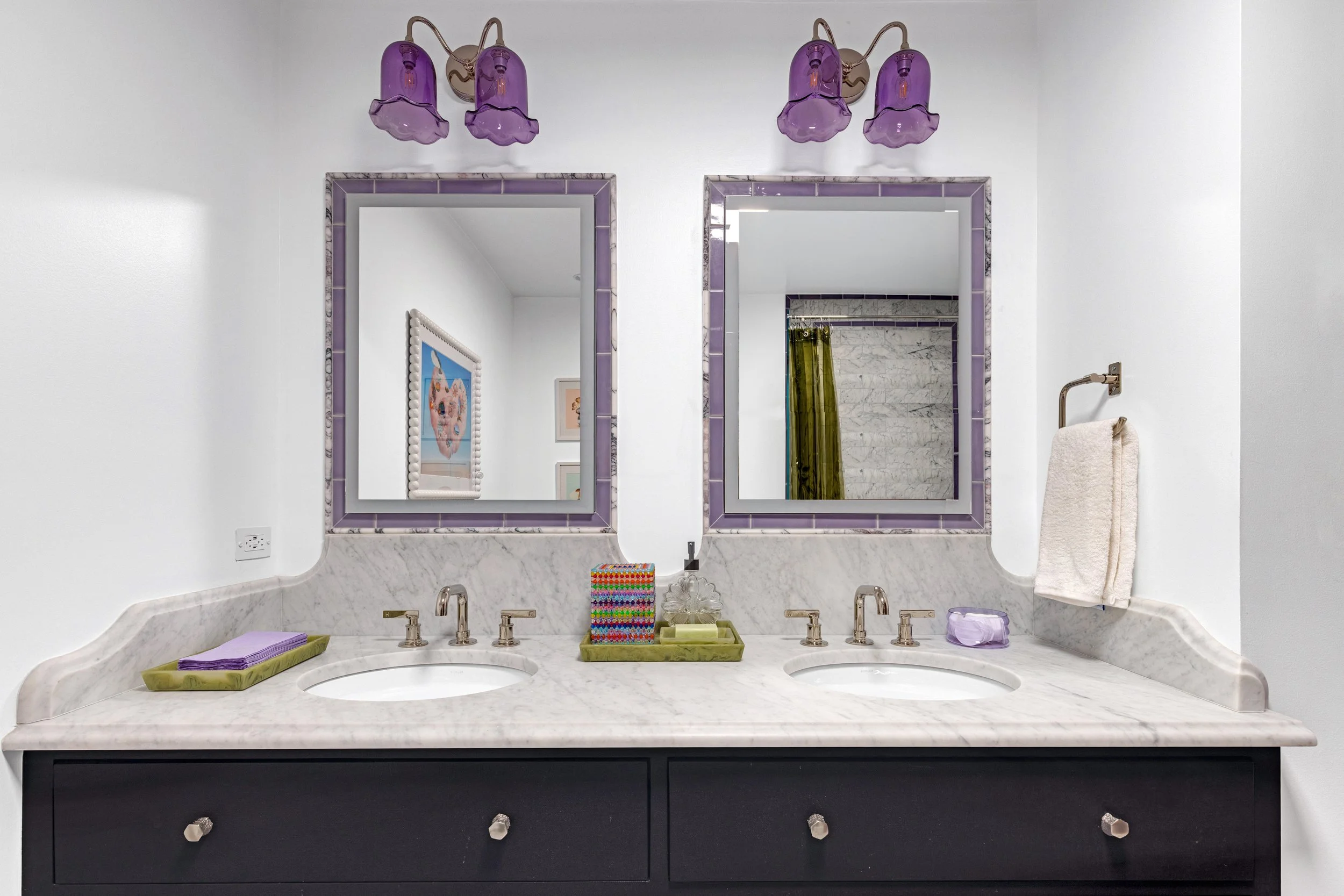 Bathroom vanity with two sinks, marble countertop, purple framed mirrors, purple and clear glass light fixtures, and various decorative items on the counter.
