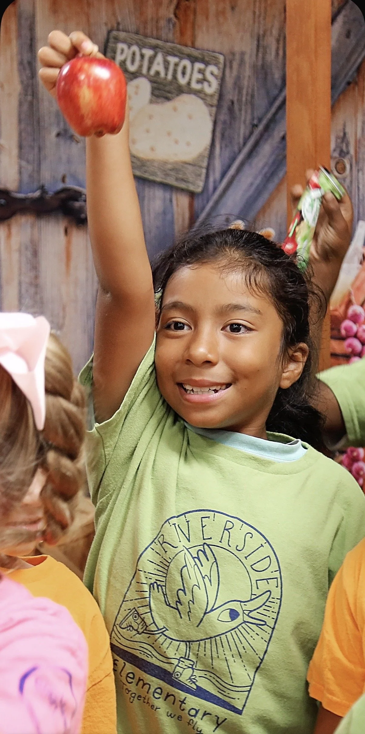 Young girl with dark hair smiling and raising her hand holding an apple in a rustic wooden room with a sign that says 'POTATOES' and other children nearby.