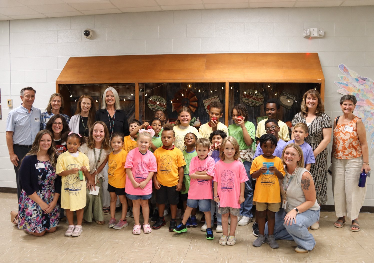 Group of children and adults posing for a photo indoors, some children wearing colorful t-shirts, some holding apples, in front of a wooden display case decorated with lights and signs, in a classroom or community center setting.