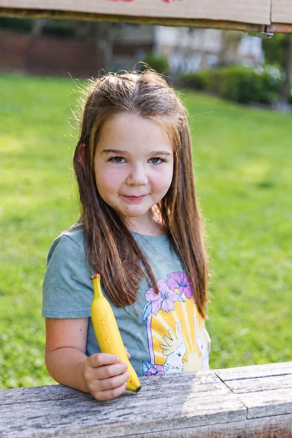 A girl with long brown hair holding a banana at an outdoor wooden table.
