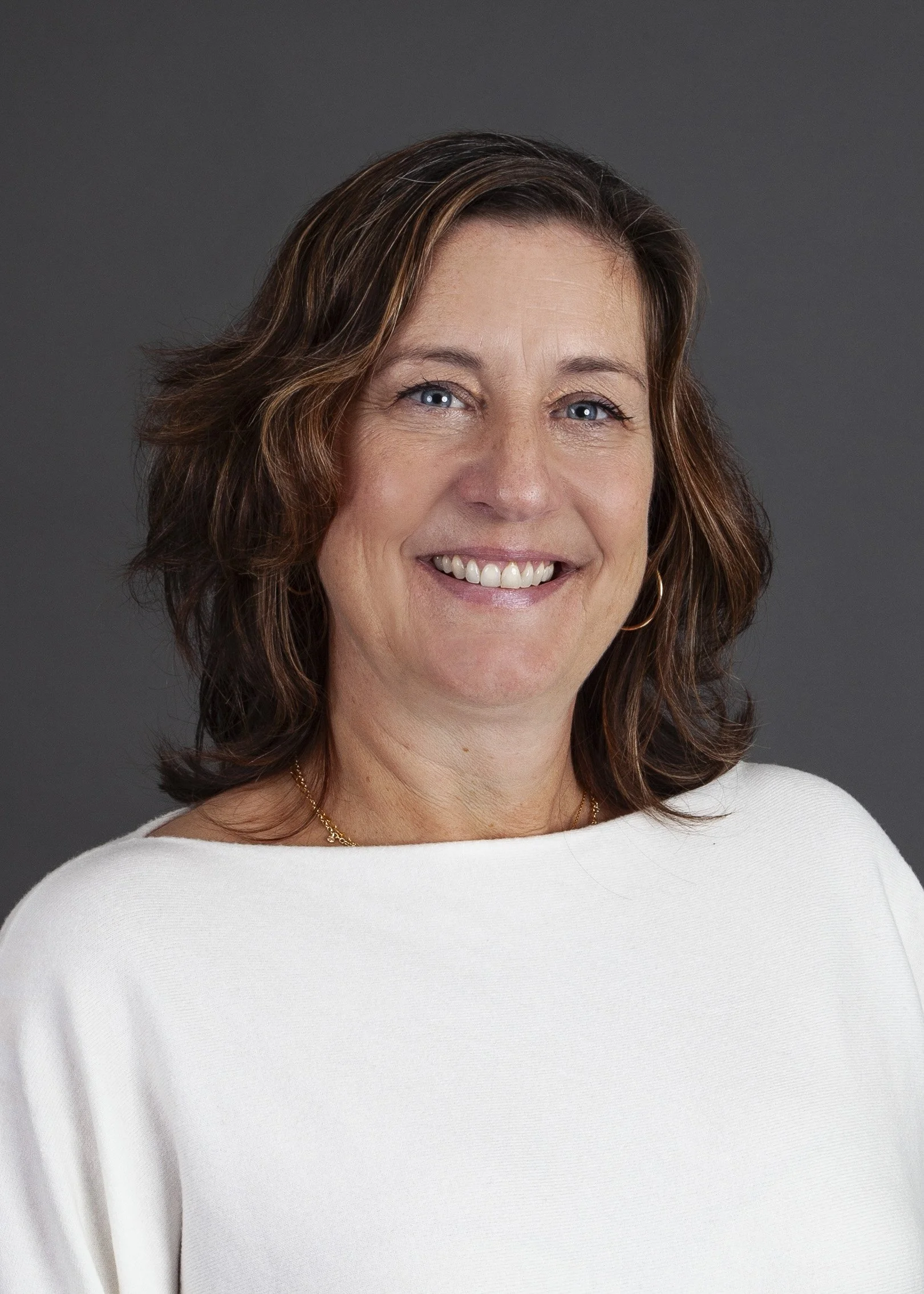 A woman with shoulder-length brown hair, smiling, wearing a white top and a gold necklace, against a dark gray background.