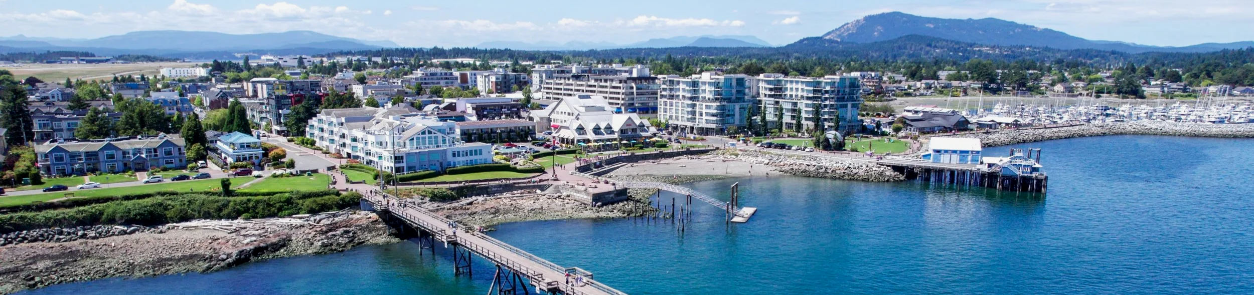 A coastal town with modern buildings, a pier extending into the water, a marina with boats, green lawns, and mountains in the background.