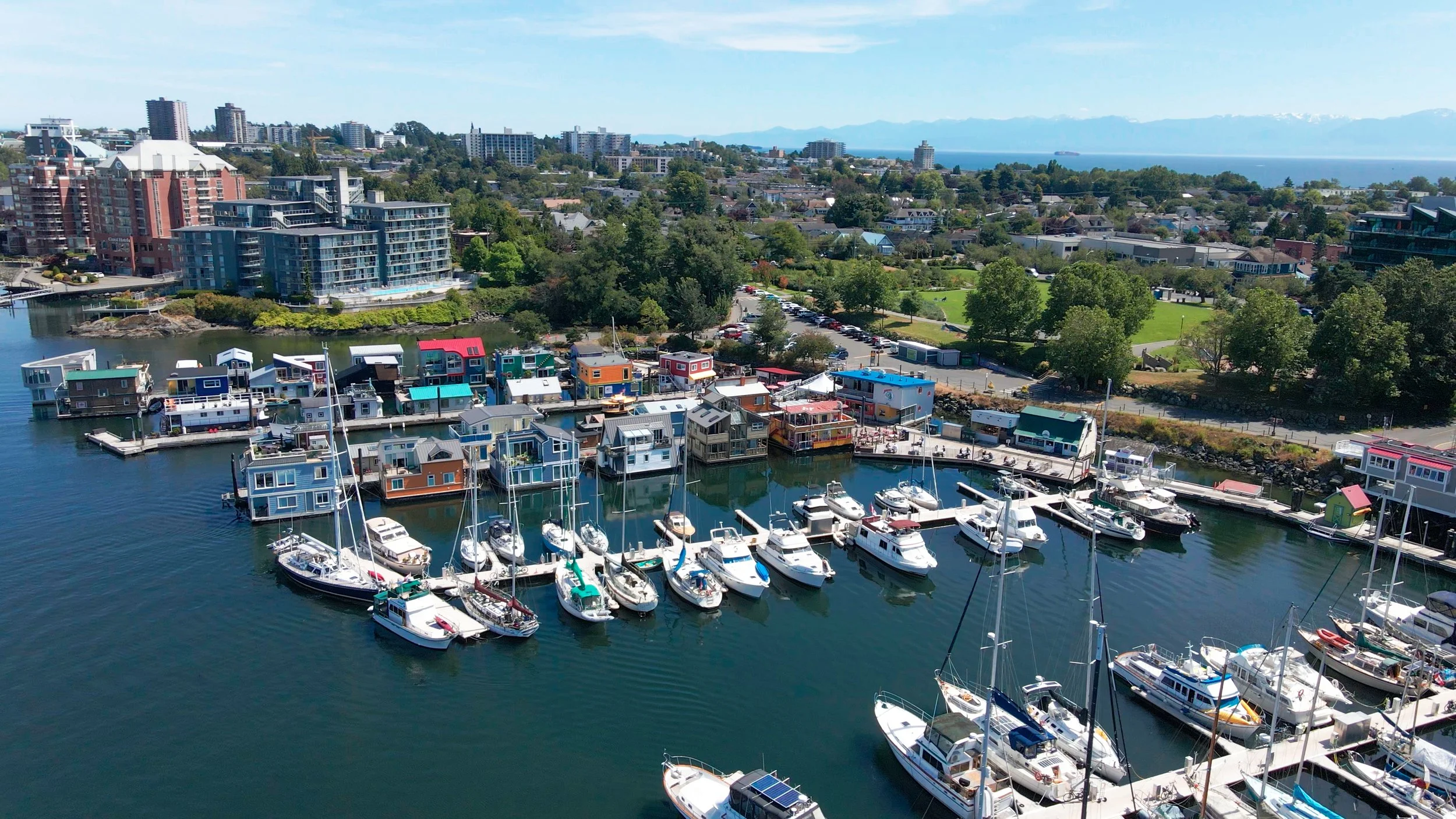 Aerial view of a marina with boats docked, colorful floating houses, a cityscape with high-rise buildings, trees, and mountains in the background.
