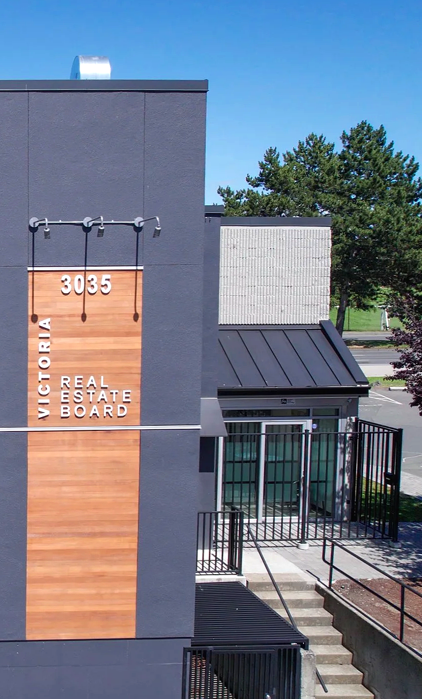 Exterior view of the Victoria Real Estate Board office building, with a gray facade, a wooden accent panel displaying the address 3035, and an entrance with steps and black railings. Trees and parking lot are visible in the background.