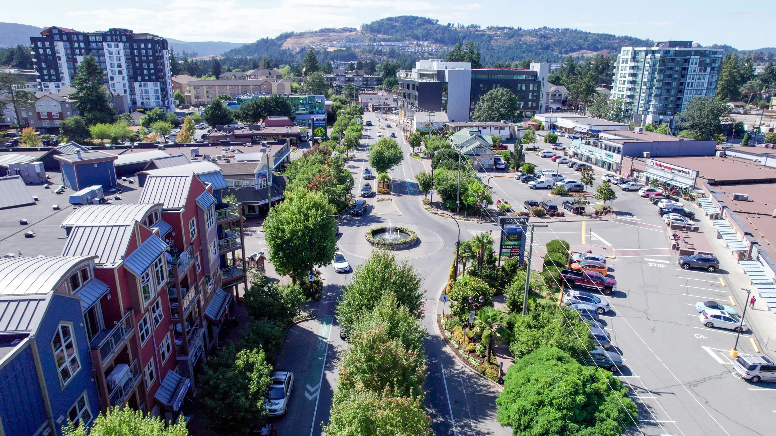 An aerial view of a small downtown area with a street running through the center, lined with trees, parked cars, and a roundabout with a fountain. Buildings, including apartments, shops, and a supermarket, surround the street, with larger residential and commercial buildings in the background against a hilly landscape.