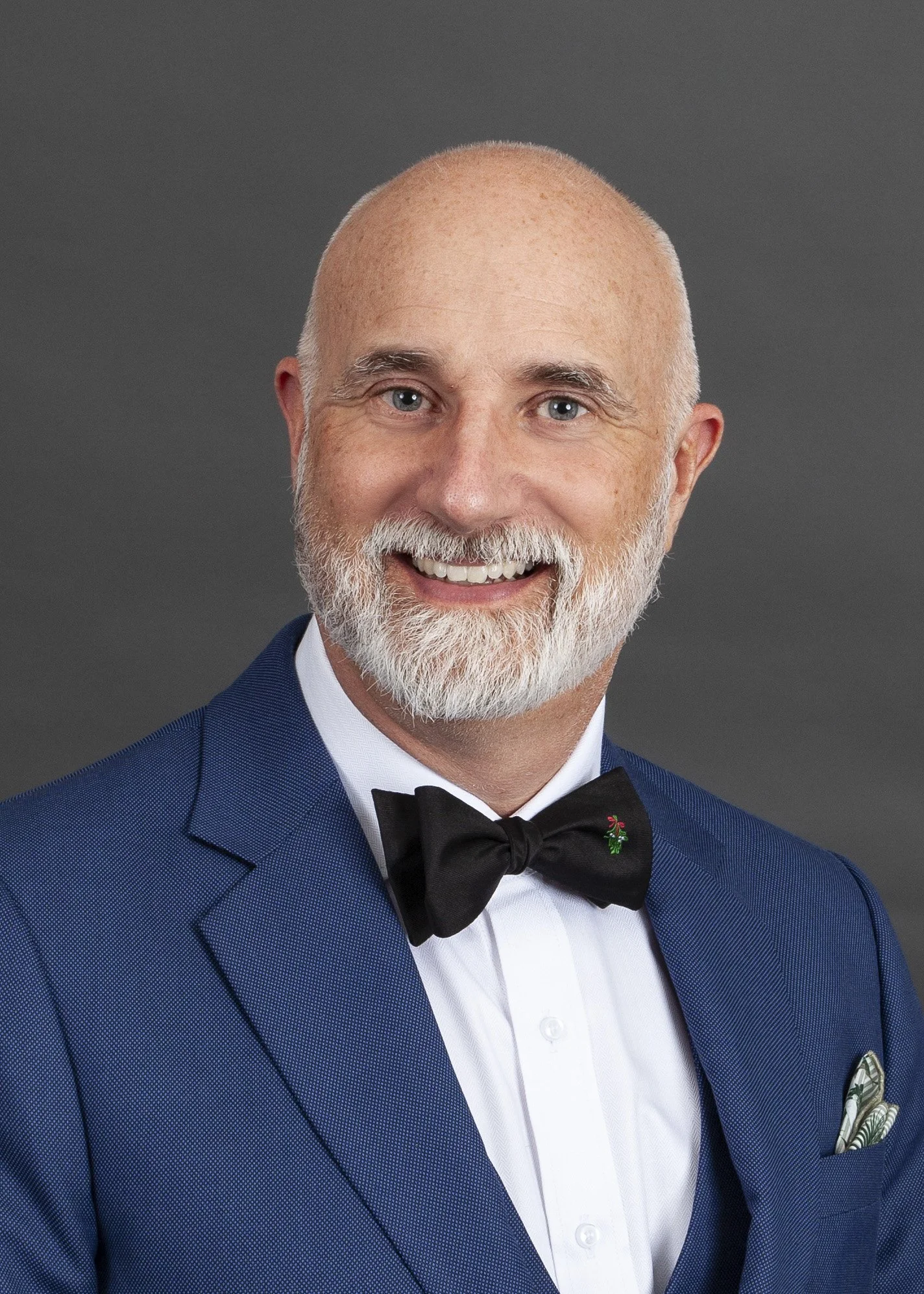 Portrait of a smiling middle-aged man with a white beard, wearing a blue tuxedo, white shirt, black bow tie, and a pocket square, against a gray background.