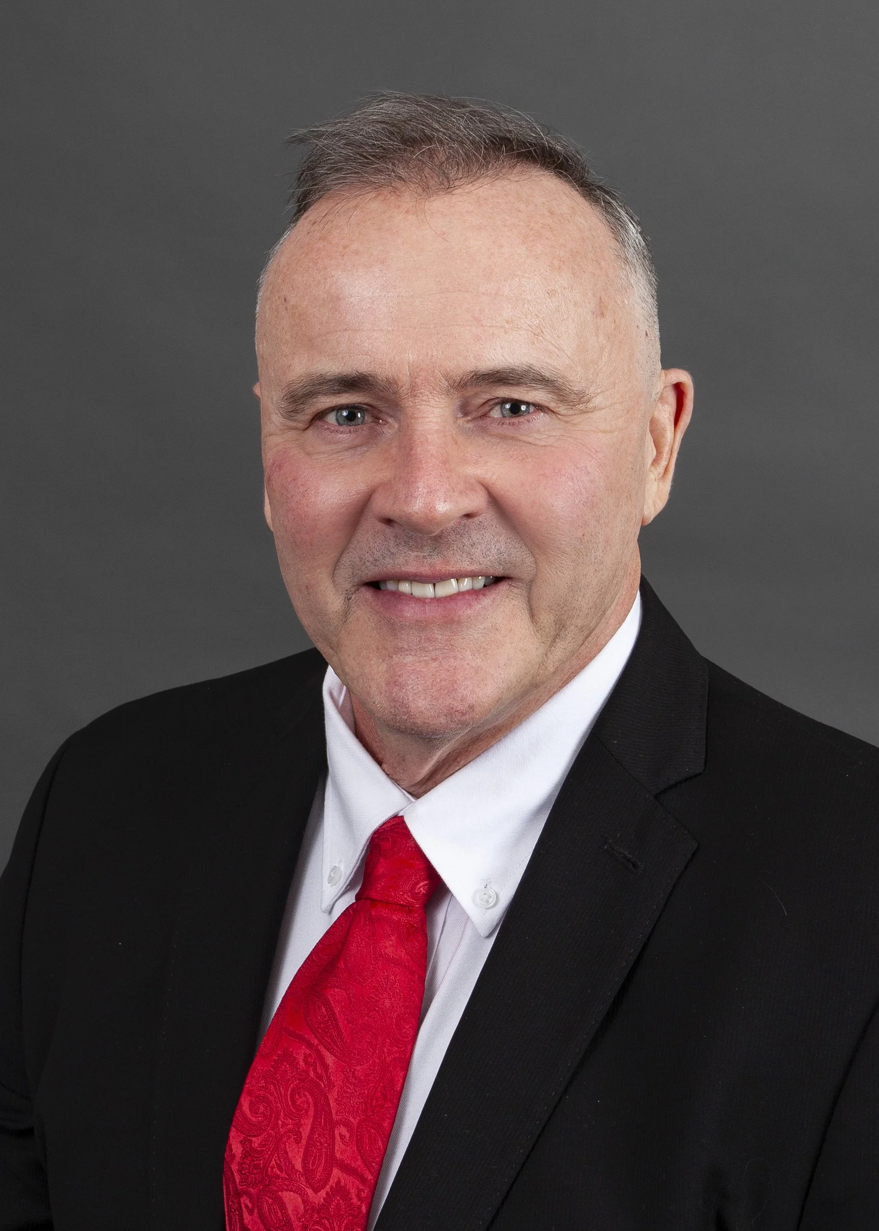 A professional man with gray hair, wearing a dark pinstriped suit and a black shirt with pink patterns, with a red poppy pinned to his lapel, smiling against a dark background.