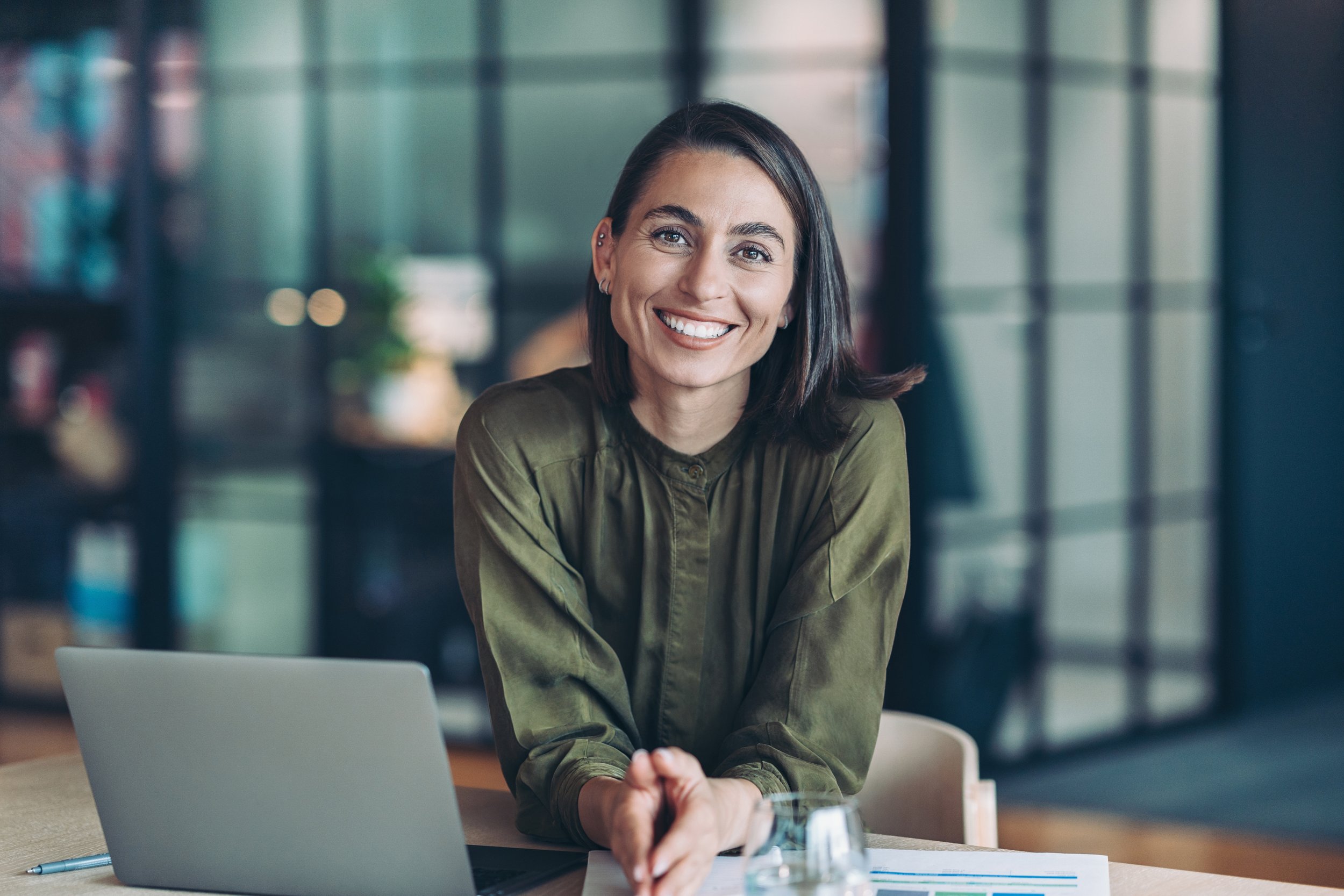 A woman with dark hair, wearing an olive green shirt, smiling and sitting at a table with a laptop and papers in a modern office setting.