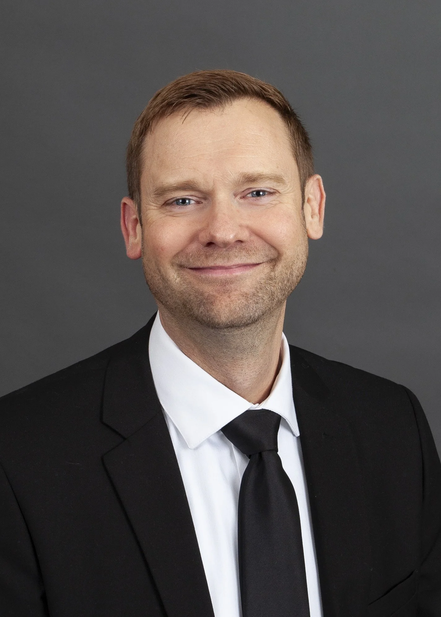 Professional headshot of a smiling man with light brown hair, wearing a dark blue suit, white shirt, and striped blue tie against a dark gray background.