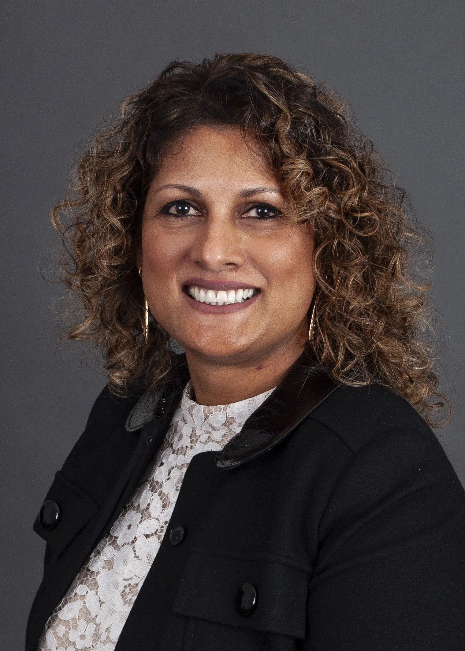 Professional headshot of a woman with curly brown hair, wearing a black blazer and a necklace with a heart-shaped pendant, smiling against a gray background.