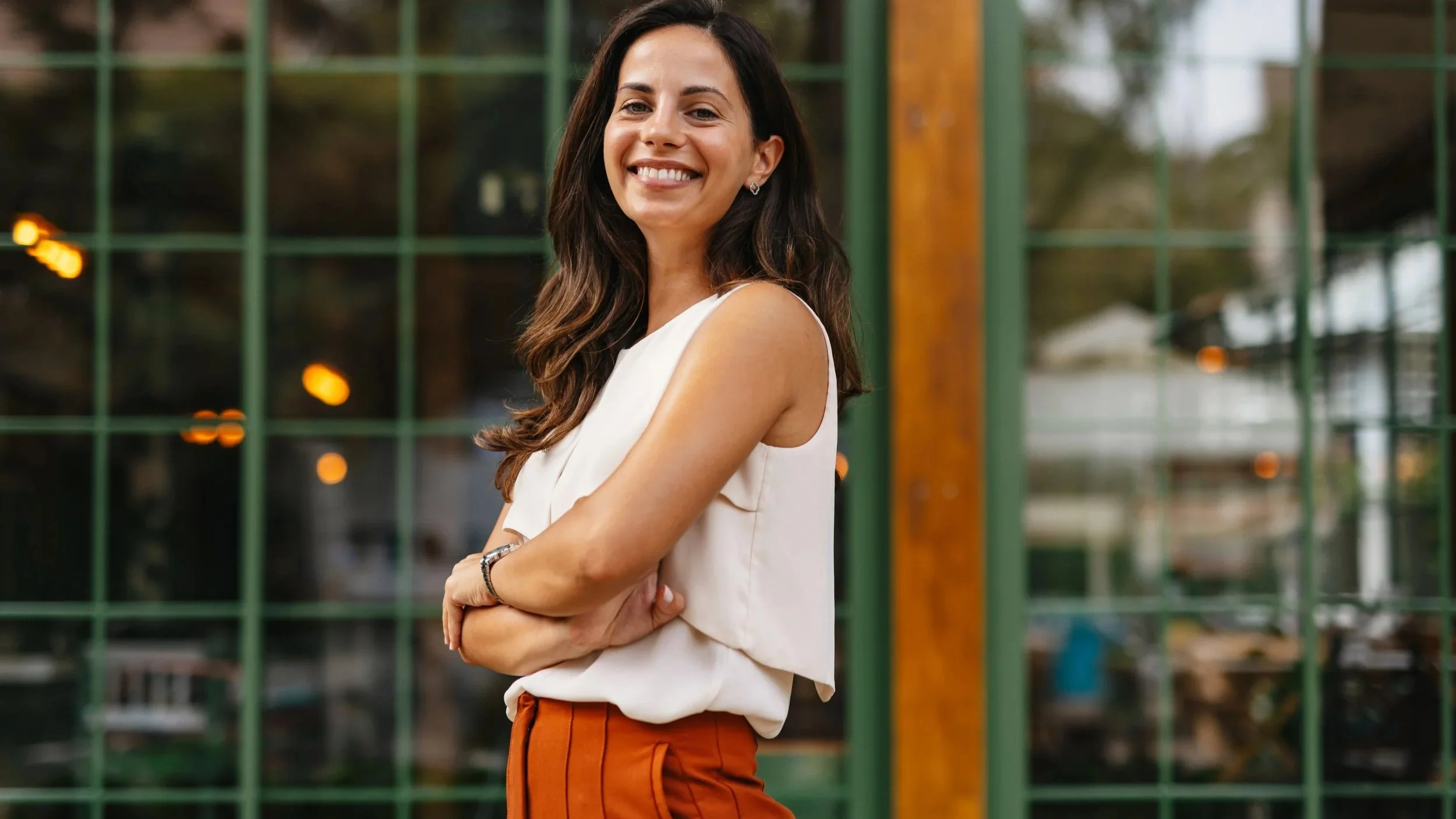 Smiling woman with crossed arms standing outside near green metal structure and glass windows.