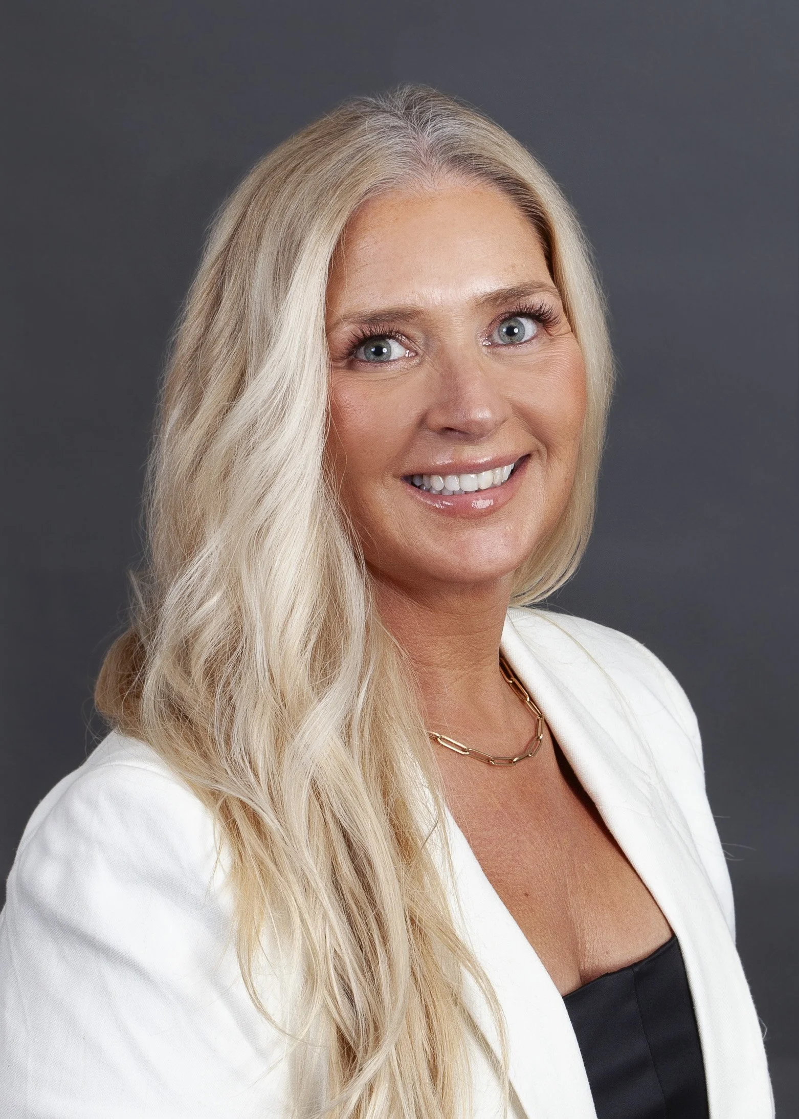 A woman with long blonde hair and blue eyes smiling, wearing a black jacket and a necklace, against a dark background.