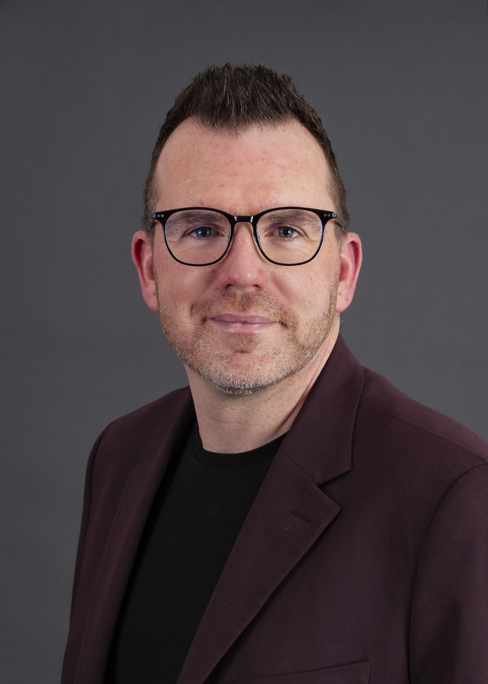 Headshot of a man with short dark hair, glasses, and a light beard, wearing a dark textured jacket against a neutral background.